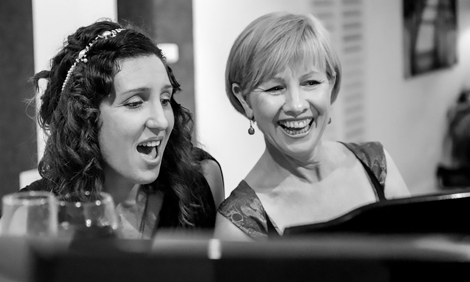 Black and white candid photo of two women at the piano singing joyfully at a community event. Photo by Raymond Mather.