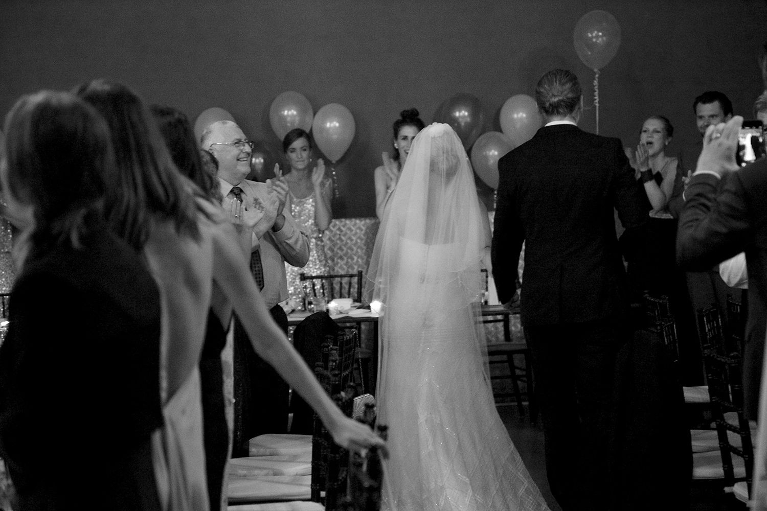 Monochrome image of newlyweds walking hand in hand through the crowd beneath balloons and soft ambient light. Photo by Raymond Mather.