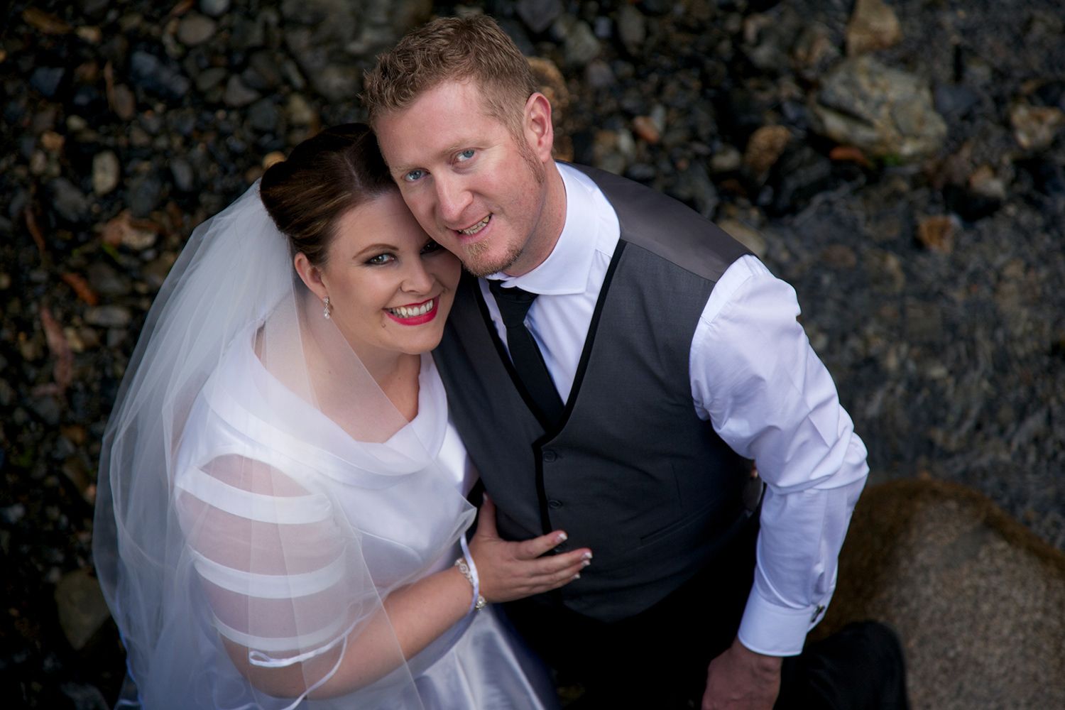 Colour portrait of newlyweds smiling up toward the camera, captured from above with natural river textures below. Photo by Raymond Mather.