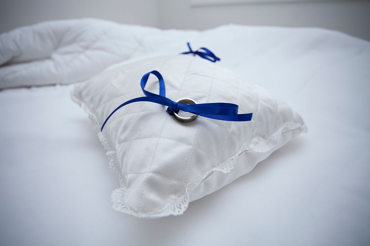 Colour detail shot of wedding rings tied with a blue ribbon on a white cushion. Photo by Raymond Mather.