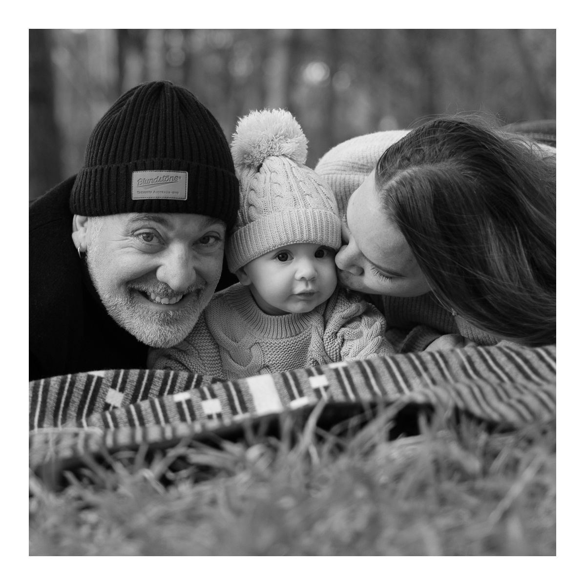 Black and white candid outdoor photo of parents kissing their baby on a picnic rug. Photo by Raymond Mather.