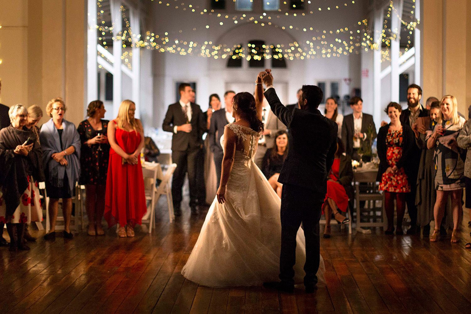 Colour photo of the couple’s first dance surrounded by family and friends under glowing fairy lights. Photo by Raymond Mather.