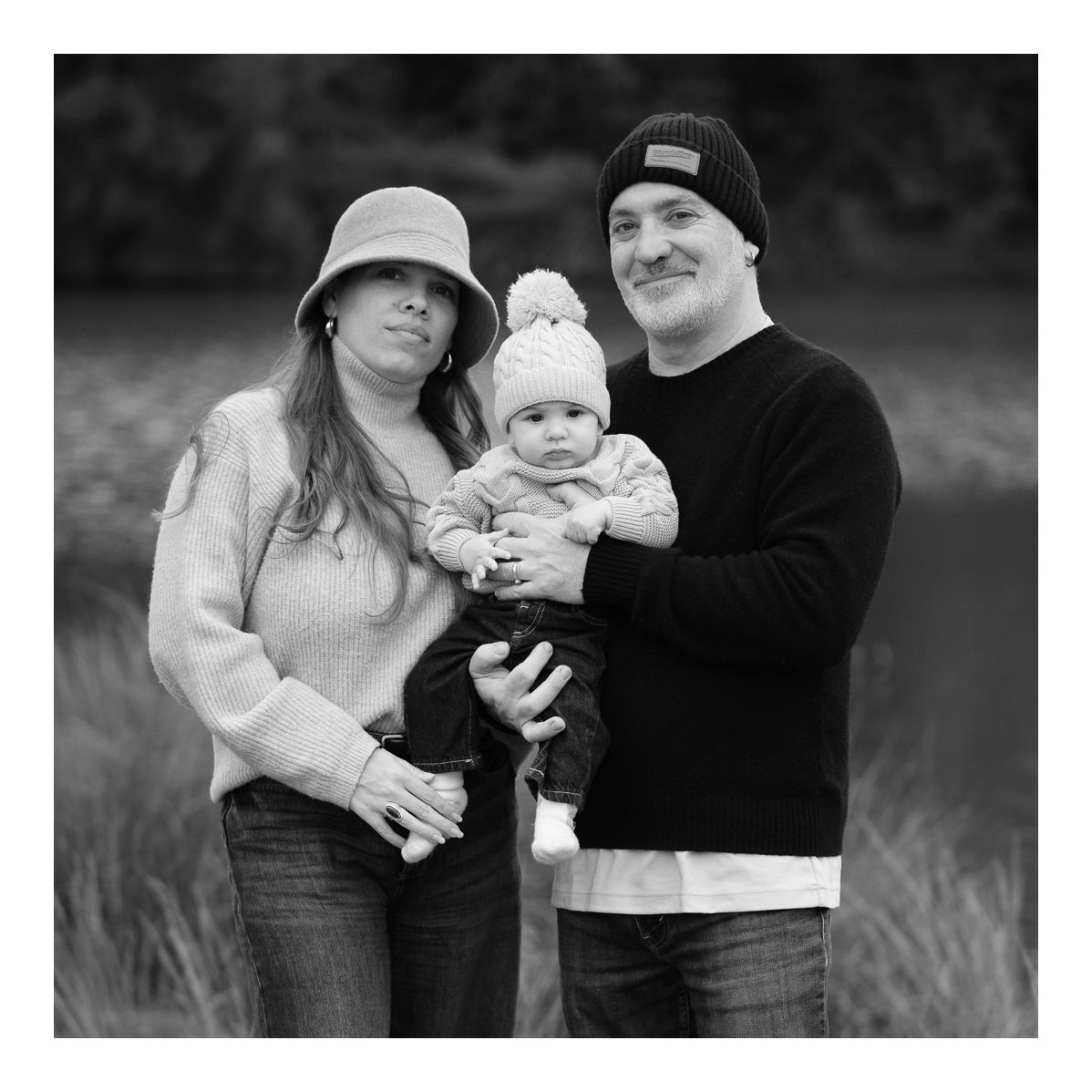 Black and white outdoor family portrait of parents standing together, smiling with baby in arms. Photo by Raymond Mather.