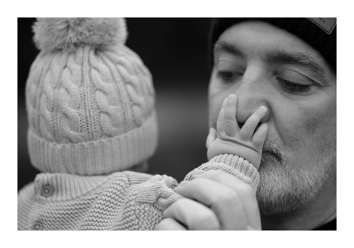 Black and white candid outdoor photo of father smiling as baby reaches for his face. Photo by Raymond Mather.