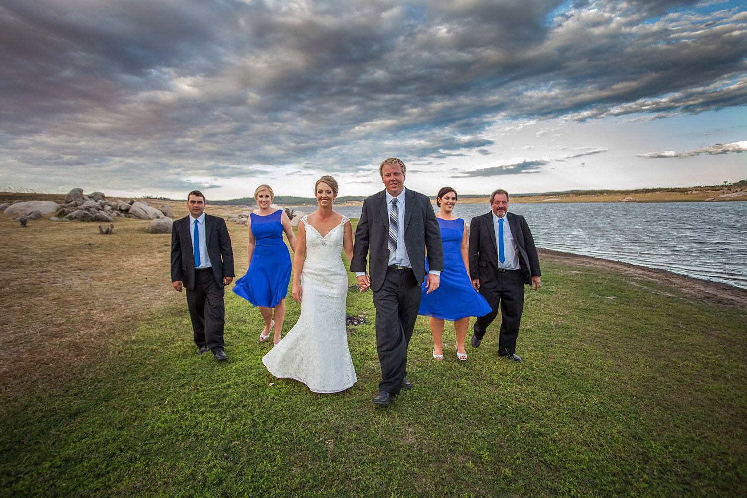 Colour wedding photo of bridal party walking along the shoreline in bright natural light. Photo by Raymond Mather.