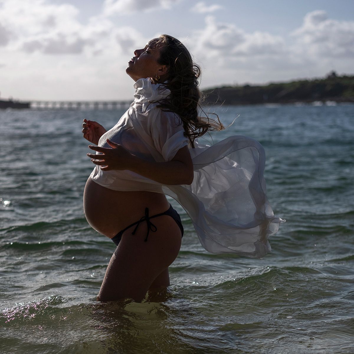 Colour outdoor maternity photo of woman standing in the sea bathed in soft afternoon light. Photo by Raymond Mather.