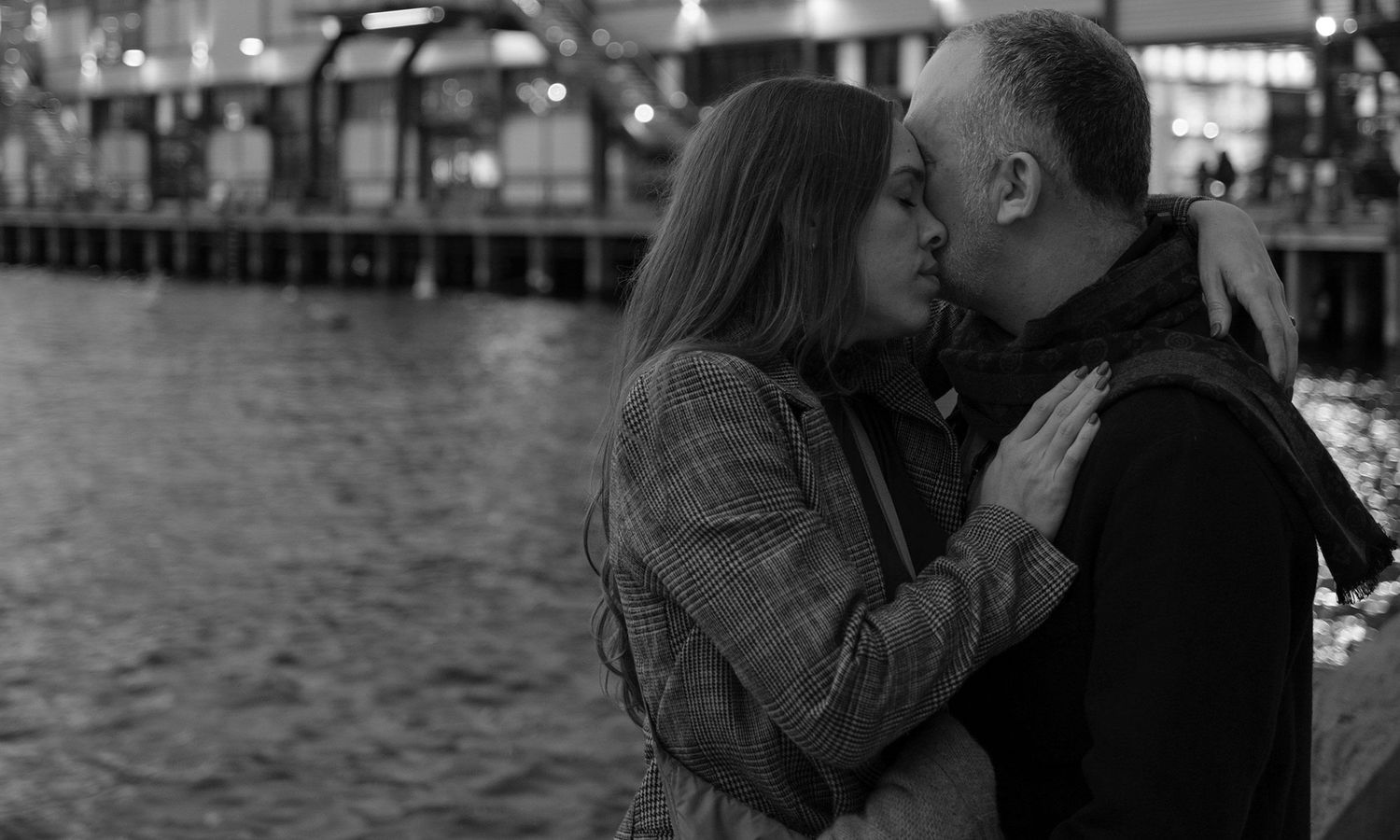 Monochrome candid photo of couple sharing a kiss beside the water at dusk. Photo by Raymond Mather.