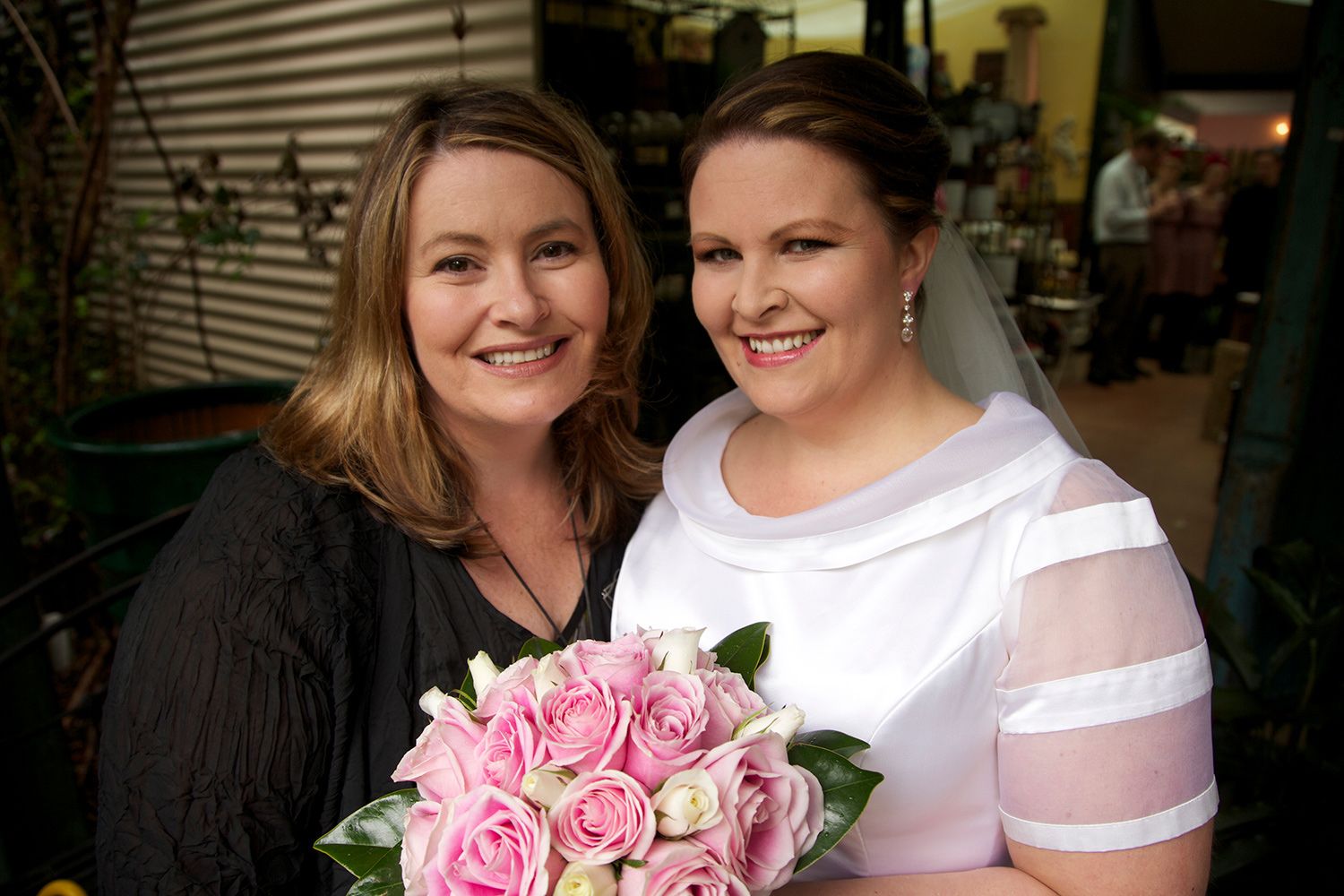 Colour portrait of bride smiling beside her mother, holding a bouquet of pink roses in warm natural light. Photo by Raymond Mather.