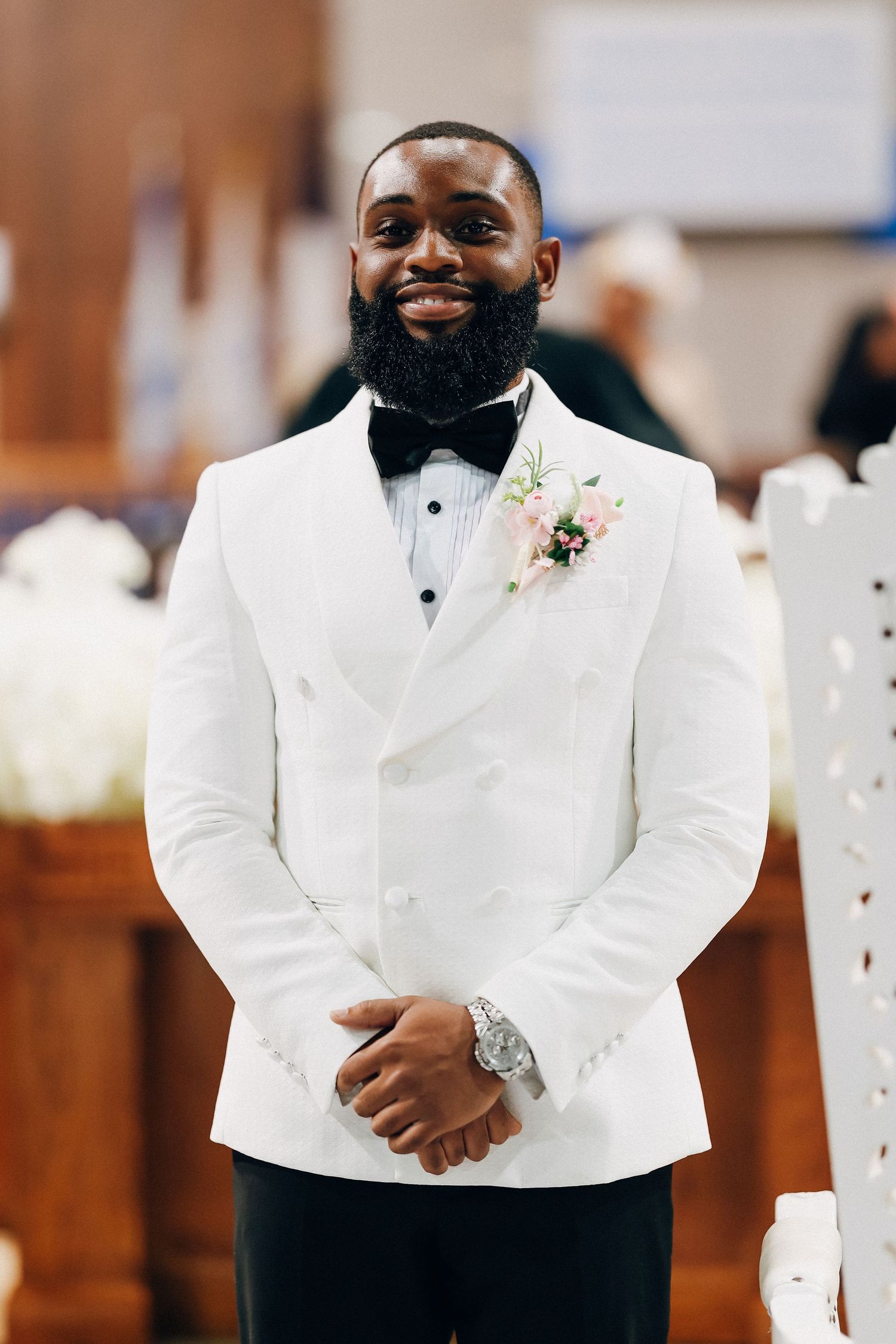 Smiling groom in white double-breasted tuxedo jacket with bow tie and floral boutonniere standing in church.