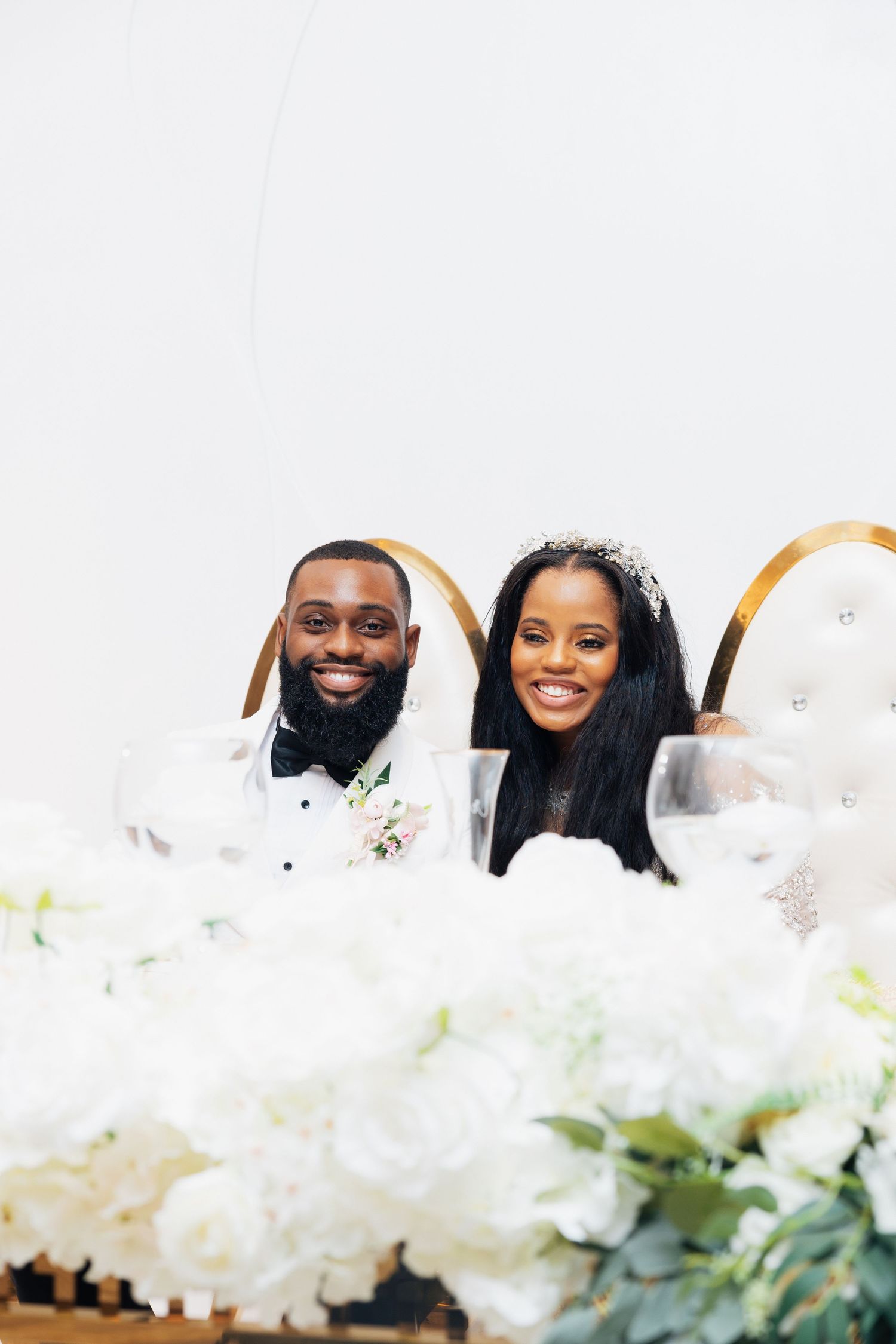 Smiling bride and groom seated at sweetheart table adorned with white floral arrangements at their wedding reception.