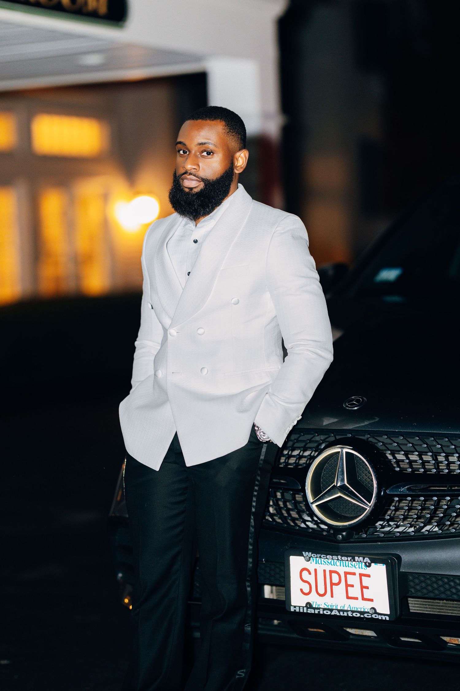 Dapper groom in white tuxedo jacket poses beside black Mercedes-Benz with SUPEE license plate at night.