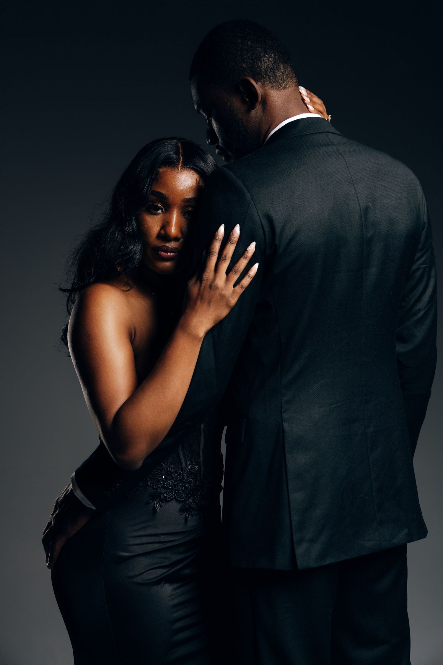 Elegant Black couple in formal attire, woman in black dress leaning on mans shoulder in dark studio.