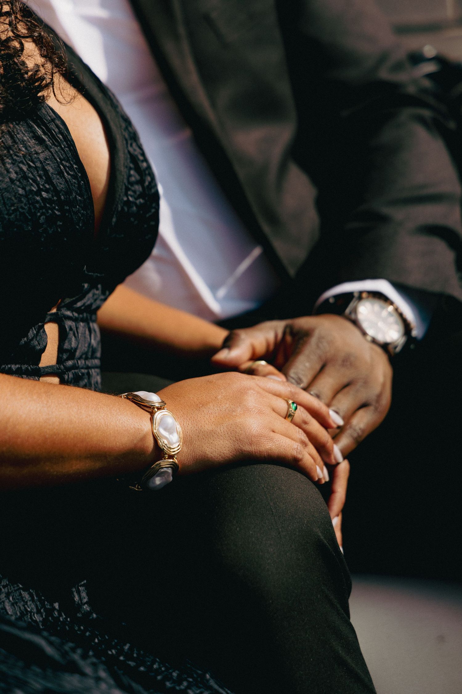 Close-up of couple holding hands, woman wearing gold bracelet and green ring, man in dark suit with silver watch.