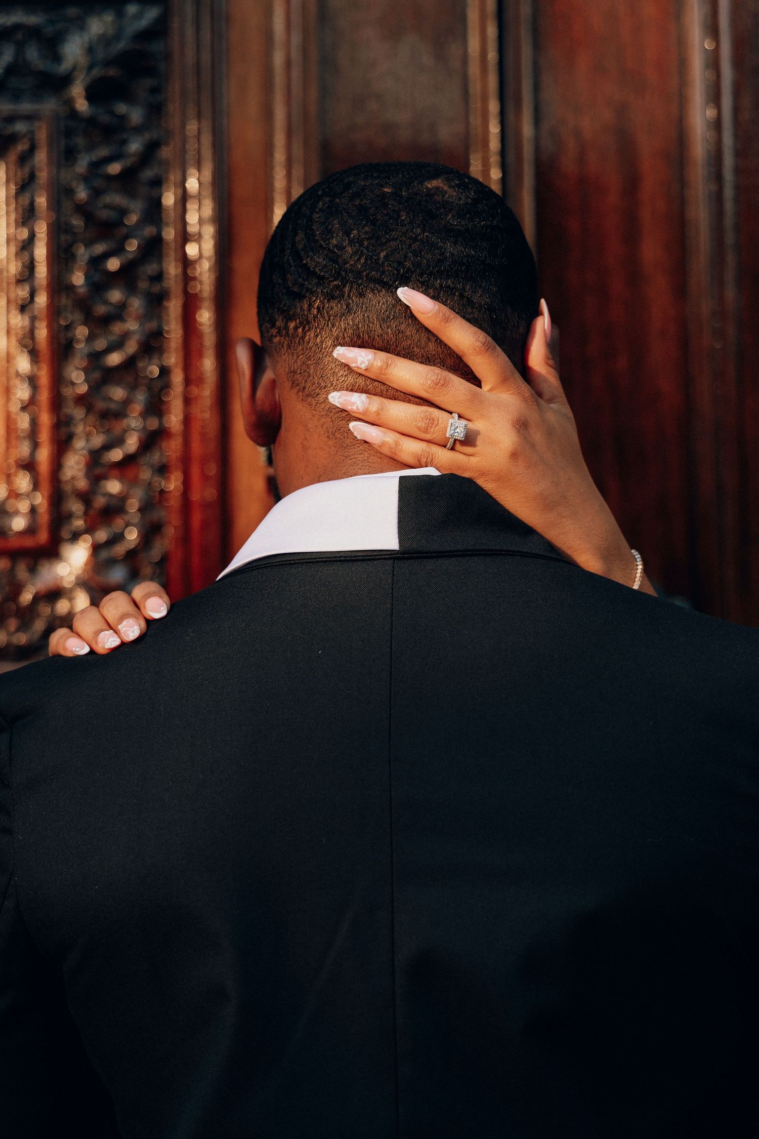 A woman's hand with an engagement ring rests on the back of a man's head, both dressed formally.