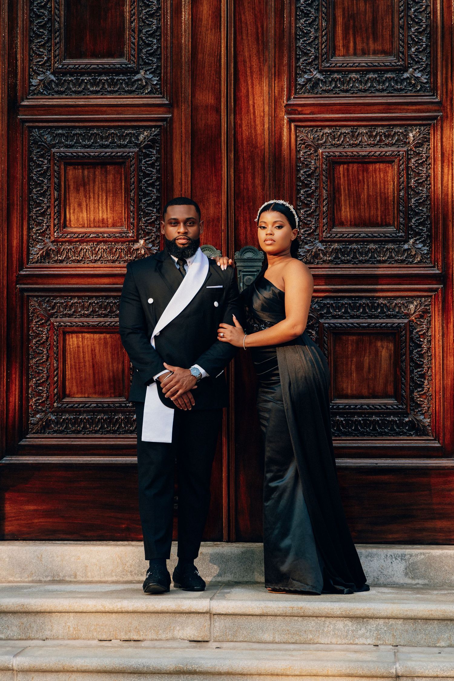 Elegant couple in black formal attire posing before ornate wooden doors, man in tuxedo and woman in sleek gown.