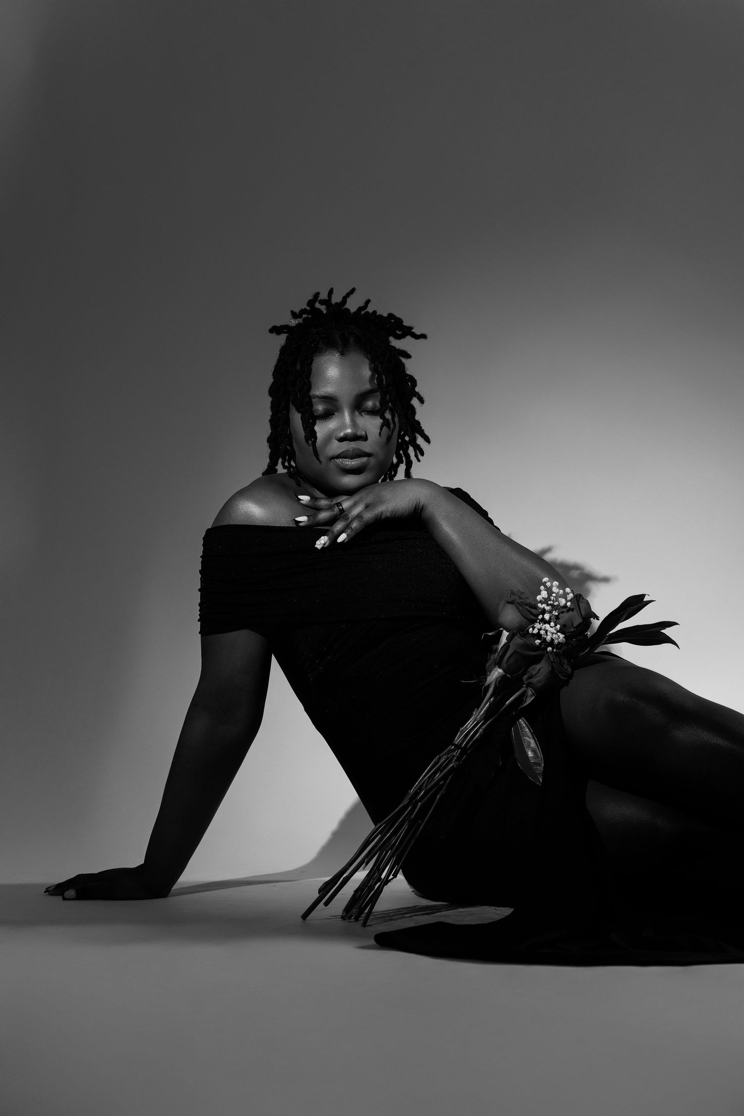 Black and white portrait of a woman with dreadlocks sitting on floor holding decorative flowers in elegant black outfit.