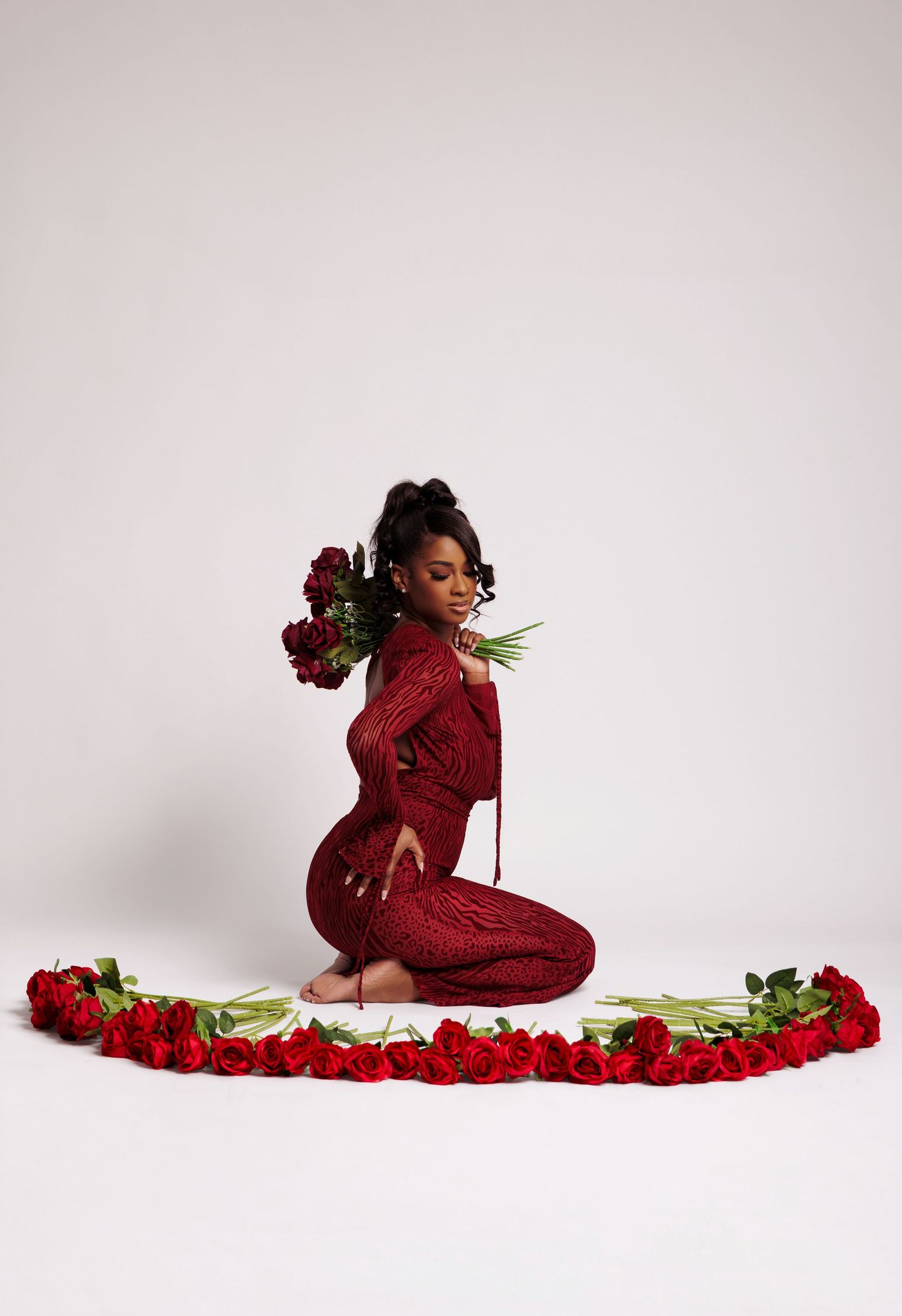 Woman in red sequin jumpsuit sits among red roses arranged in a semicircle, holding bouquets, on white background.