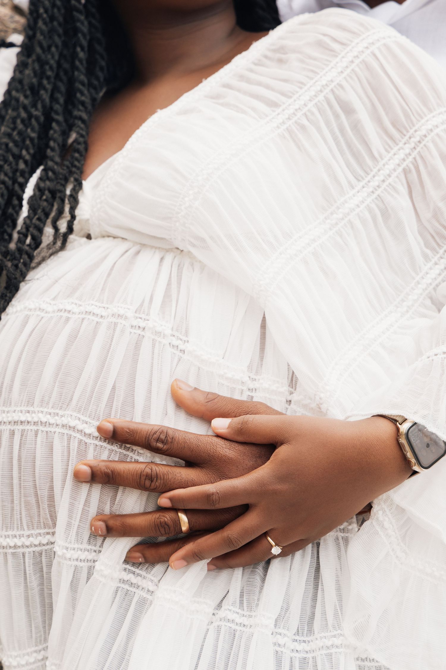 Pregnant woman in white ruffled dress with partner's hands gently cradling her baby bump, both wearing gold rings.