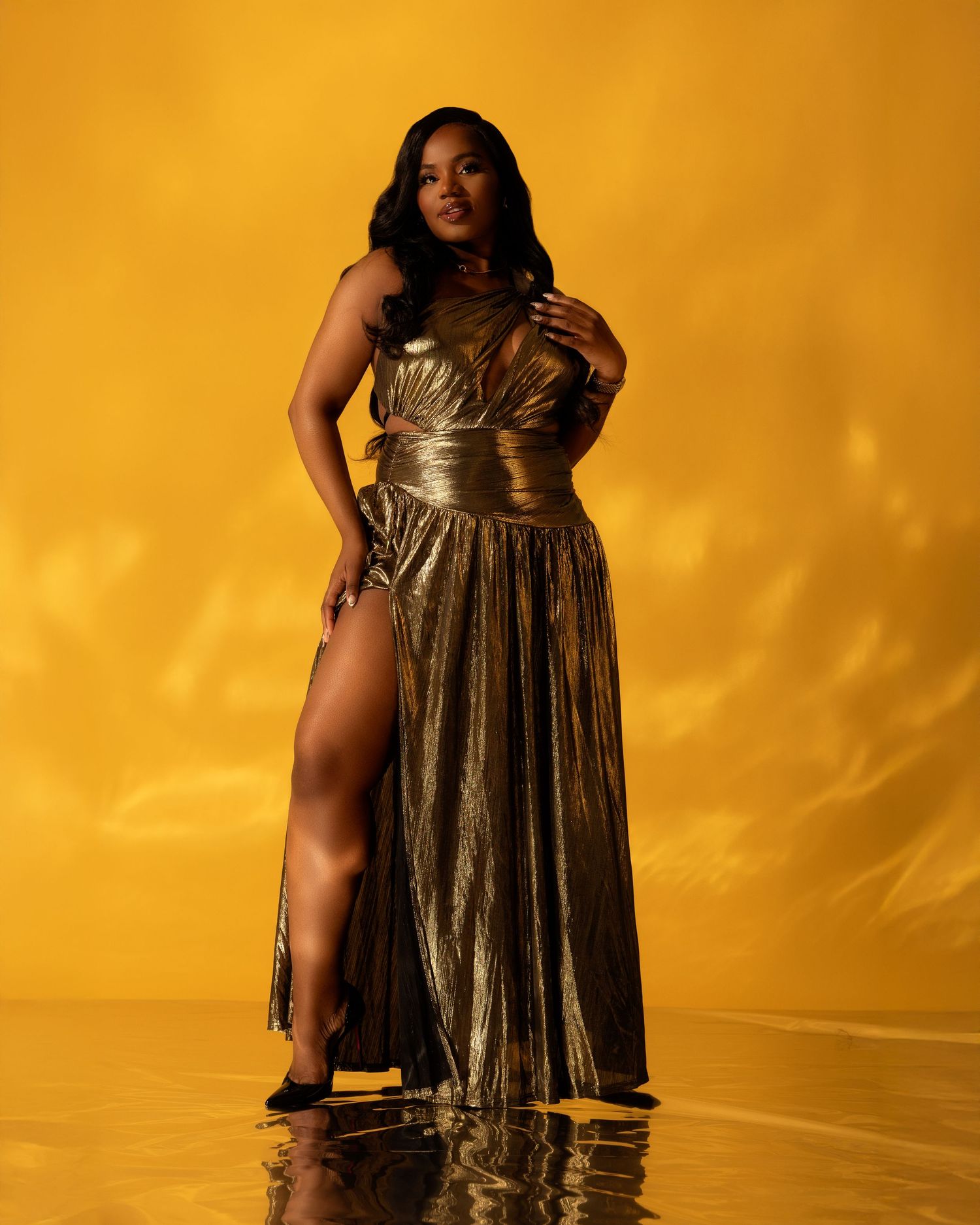 Woman in gold metallic gown poses elegantly against a golden yellow backdrop in a glamorous fashion portrait.