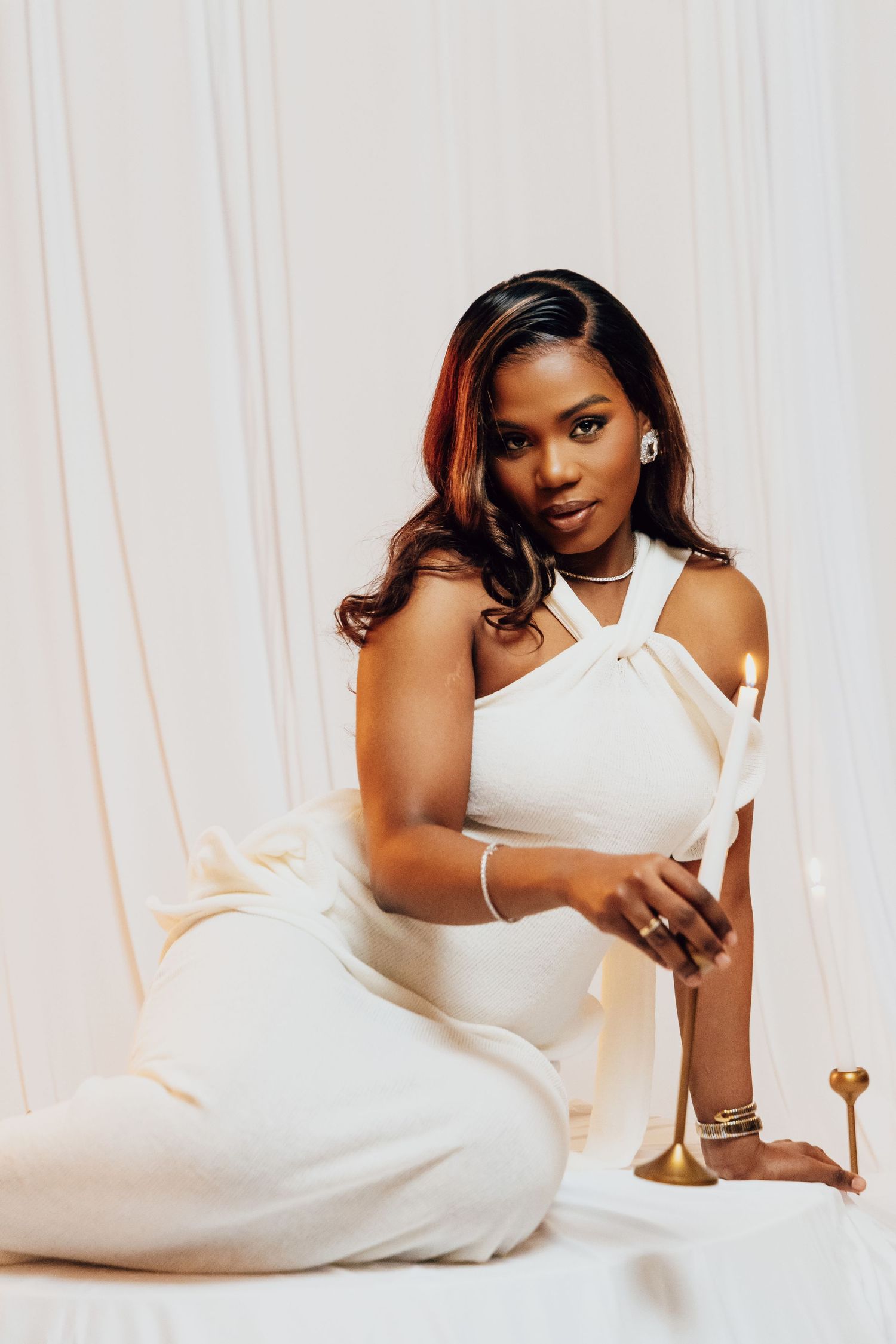 A woman in a white halter jumpsuit sits gracefully on the floor holding a gold candlestick against a white draped backdrop.