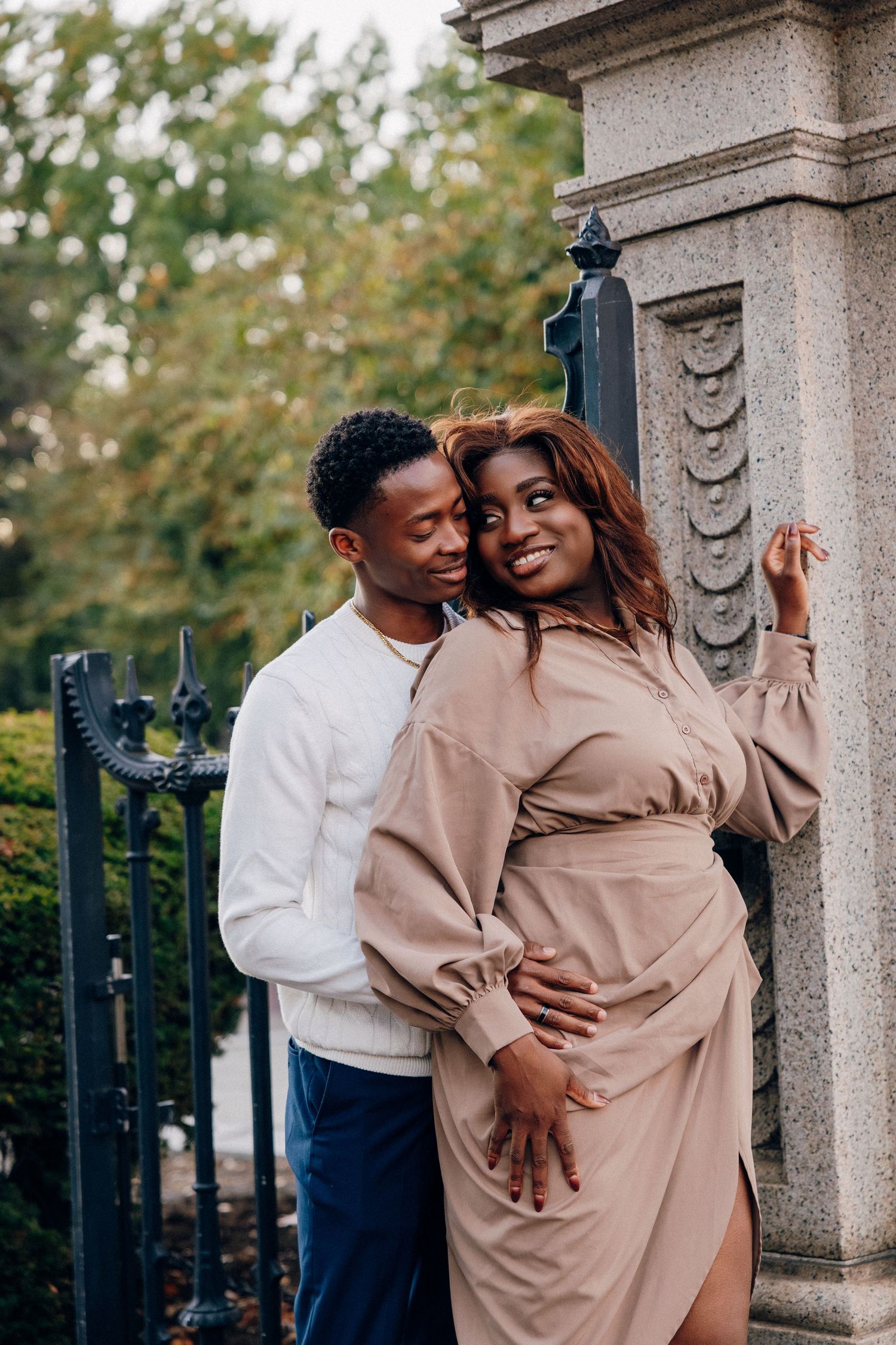 A couple embraces near ornate iron gates and stone pillars, surrounded by lush green trees outdoors.