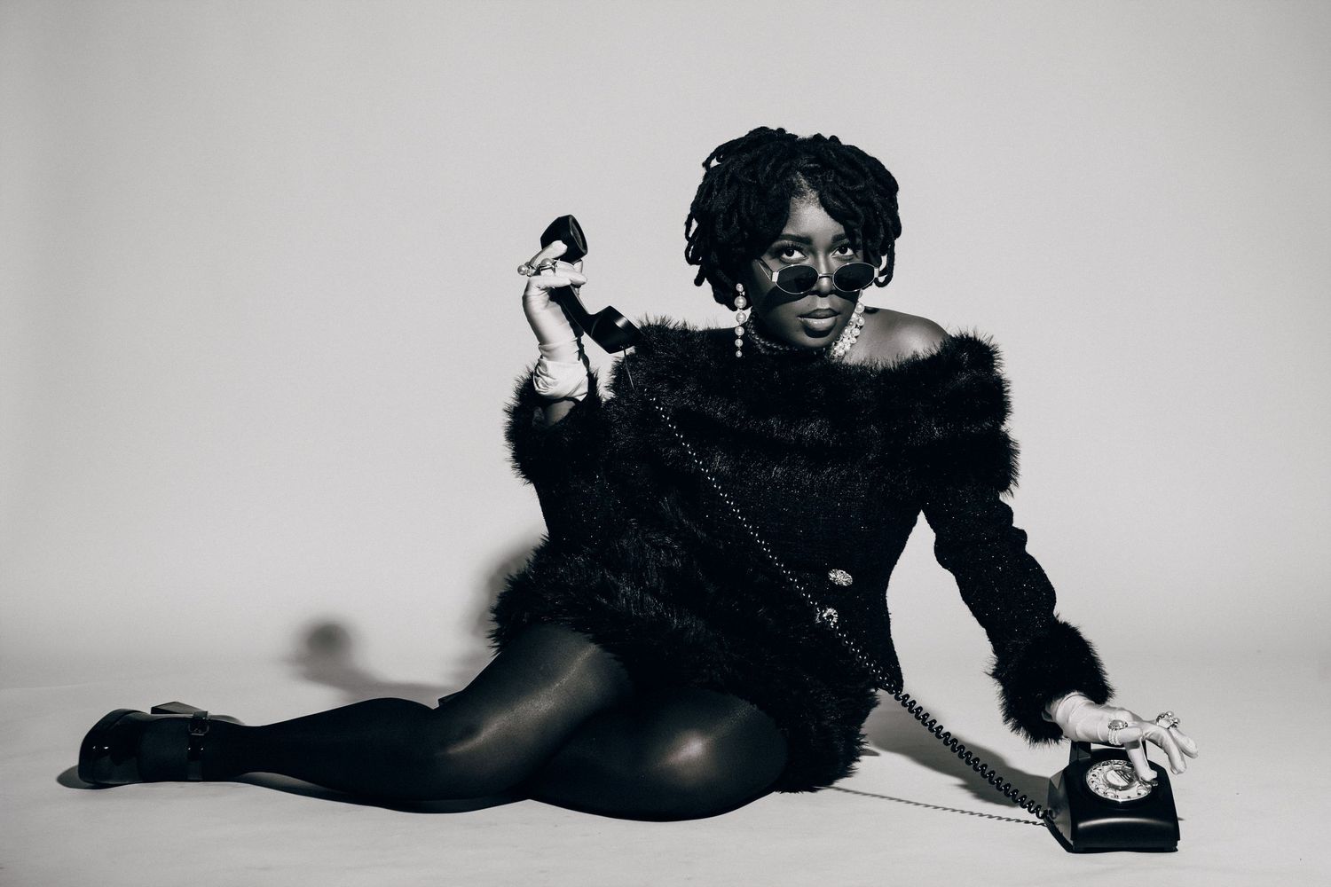 Black and white photo of a stylish woman in fur jacket and leather pants posing with a vintage telephone.