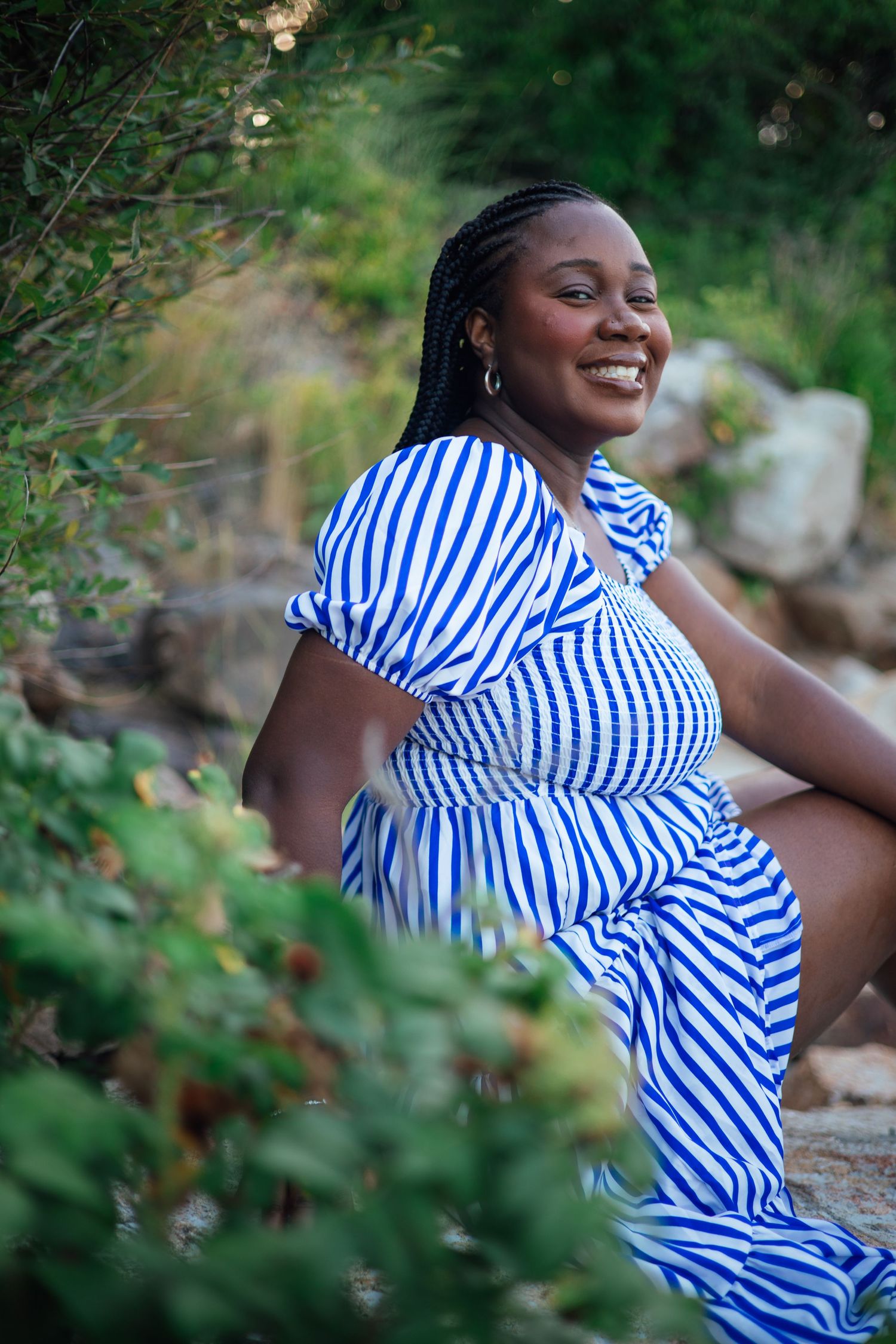 Smiling young Black woman in blue and white striped dress sitting outdoors among lush green plants and rocks.