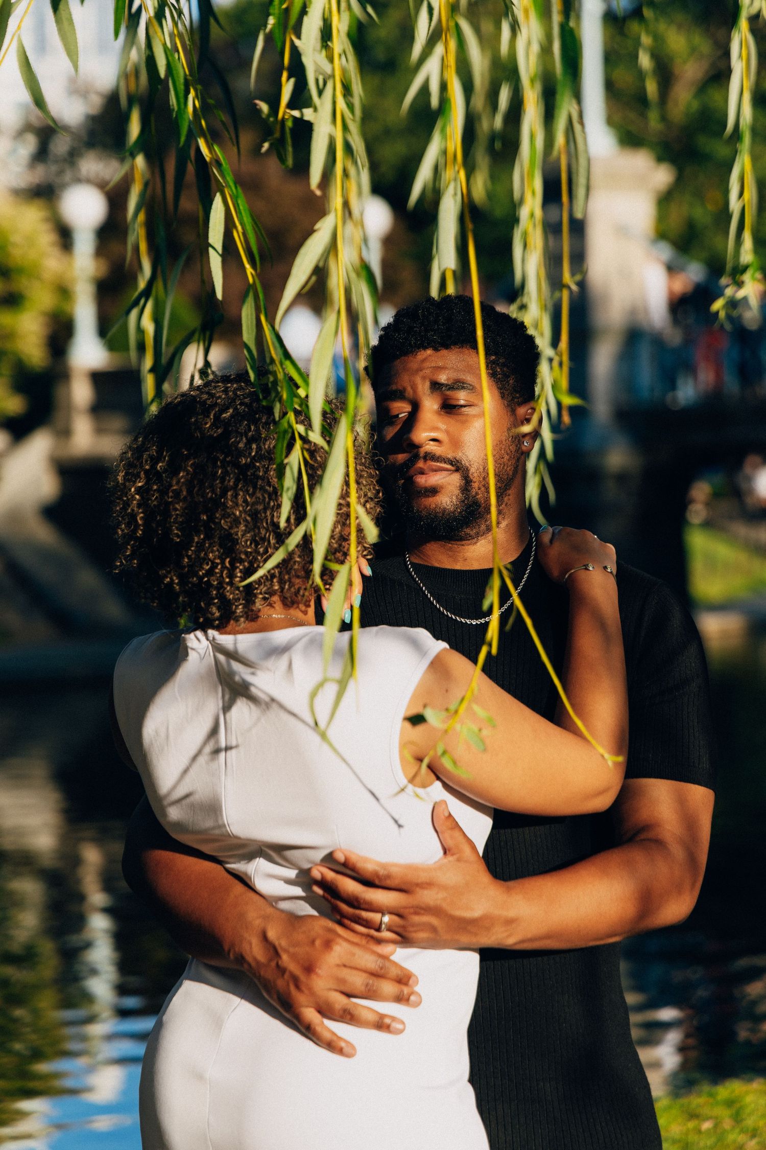 A couple embraces tenderly under weeping willow branches near a sunlit canal, woman in white dress, man in black shirt.