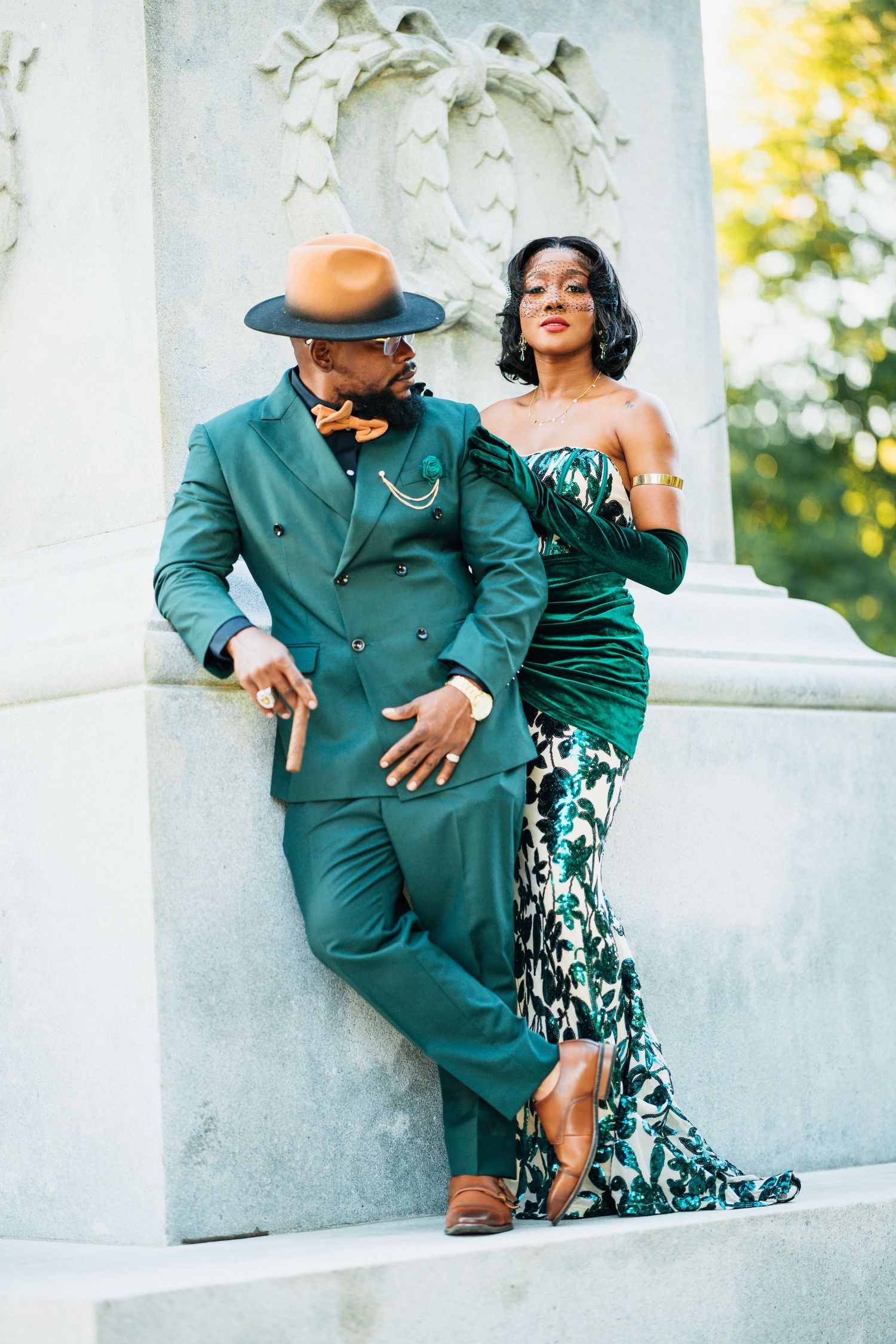 Fashionable couple posing by a stone column, man in teal double-breasted suit with hat, woman in floral gown.