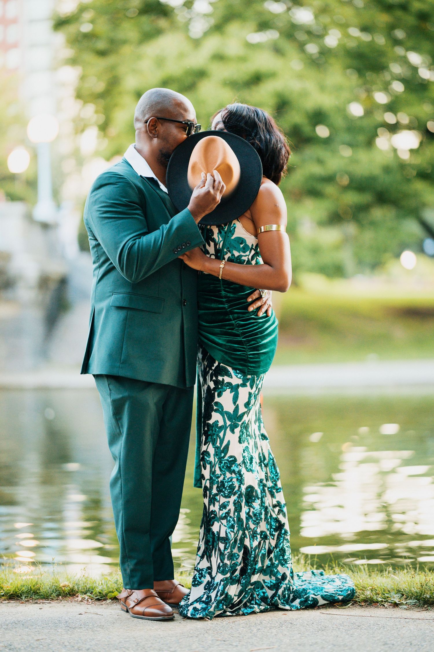 Romantic couple in matching green outfits sharing a kiss hidden behind a wide-brim hat by a reflective pond.