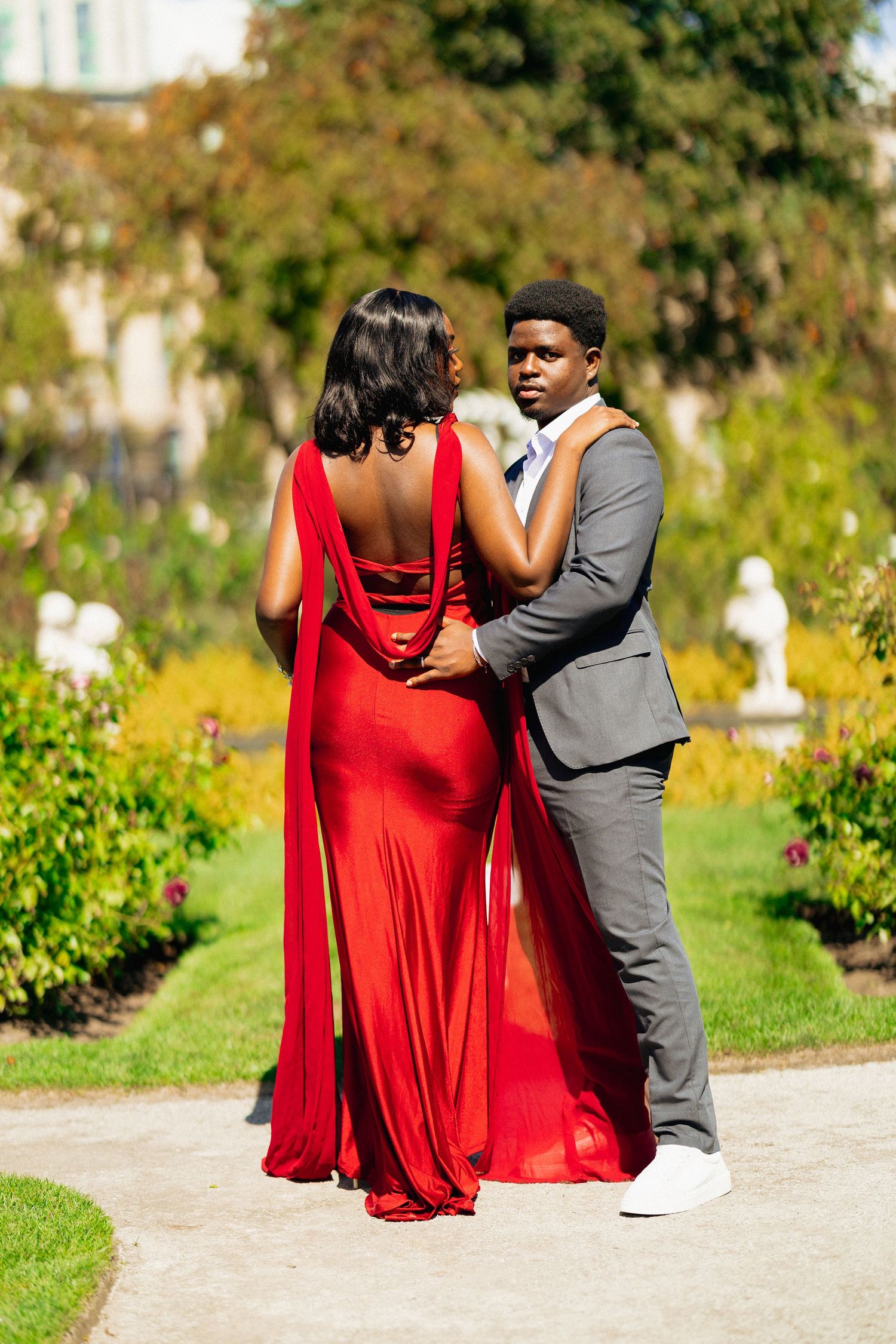 Couple embracing in sunny garden, woman in elegant red cape gown and man in gray suit.