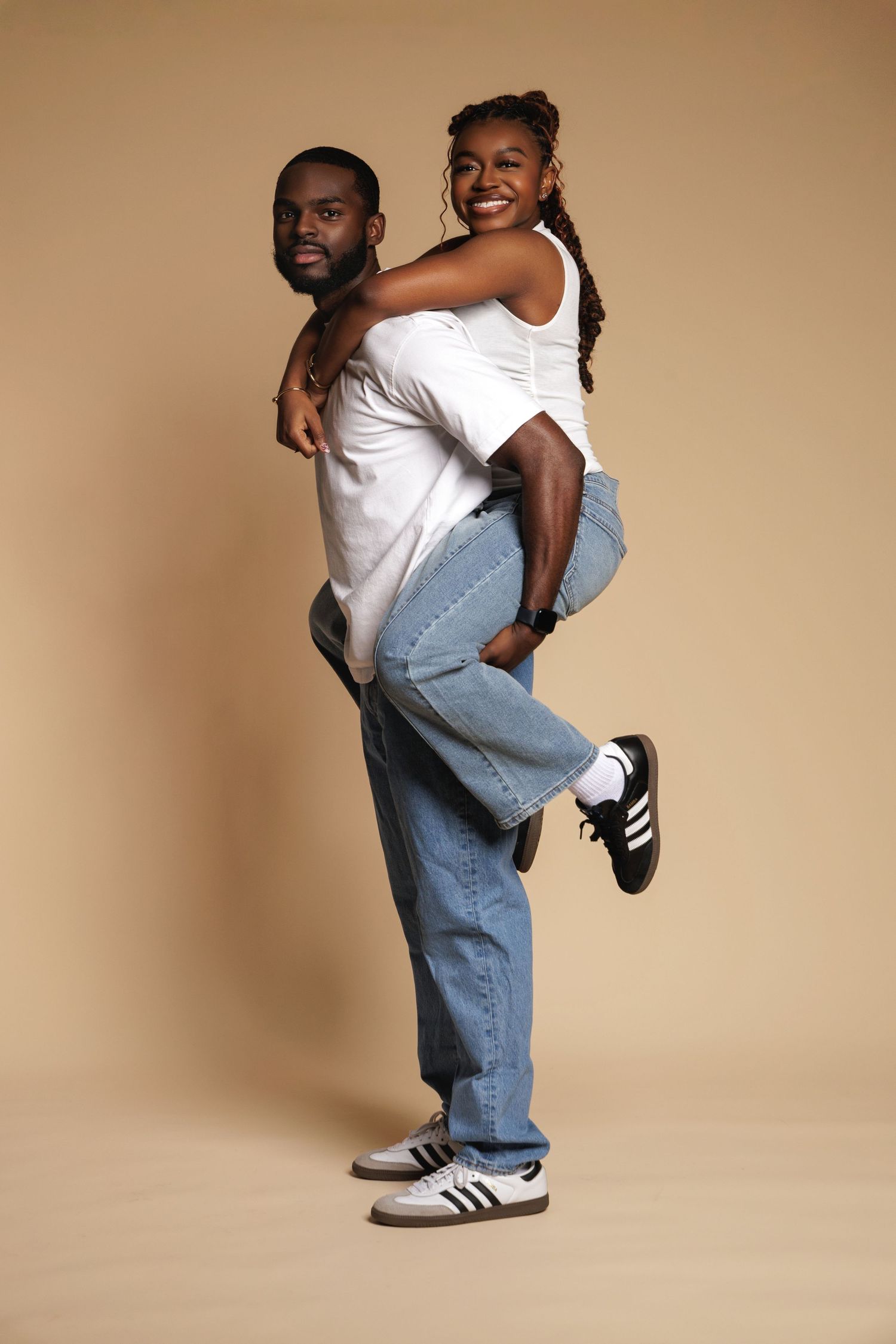 A man gives a smiling woman a piggyback ride, both wearing white tops and jeans on a beige background.