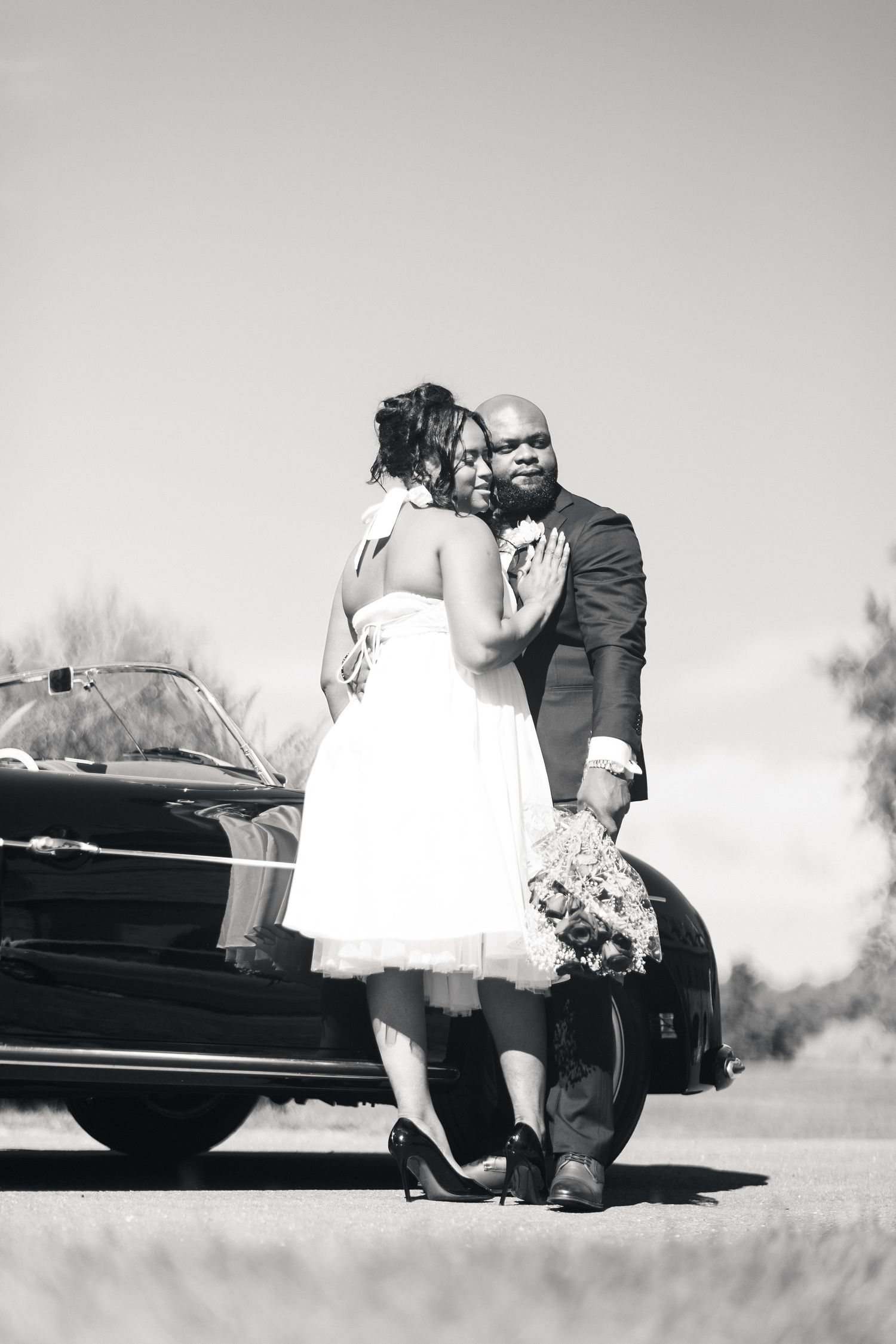 Black and white photo of bride and groom embracing beside a vintage convertible on a sunny day.