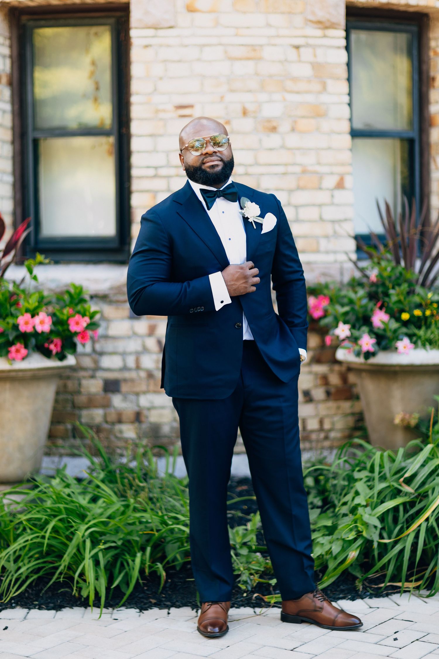 Handsome groom in navy suit with bow tie and boutonniere poses before a brick building.