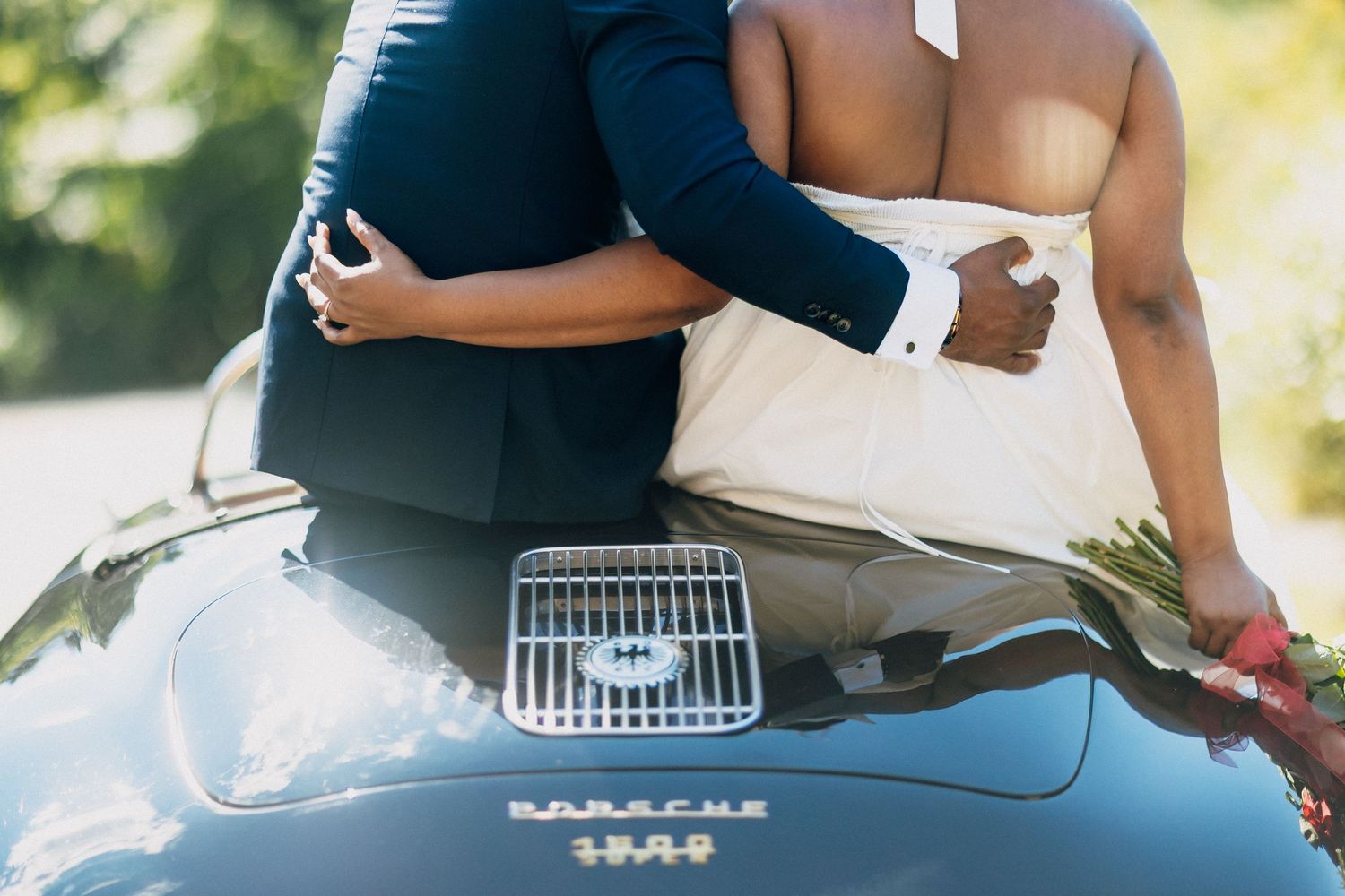 Couple embraces on the hood of a vintage Porsche 356, groom holding a red floral bouquet.