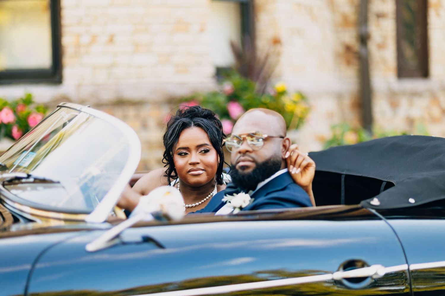 Bride and groom sit together in a classic black convertible car, smiling, surrounded by colorful garden flowers.