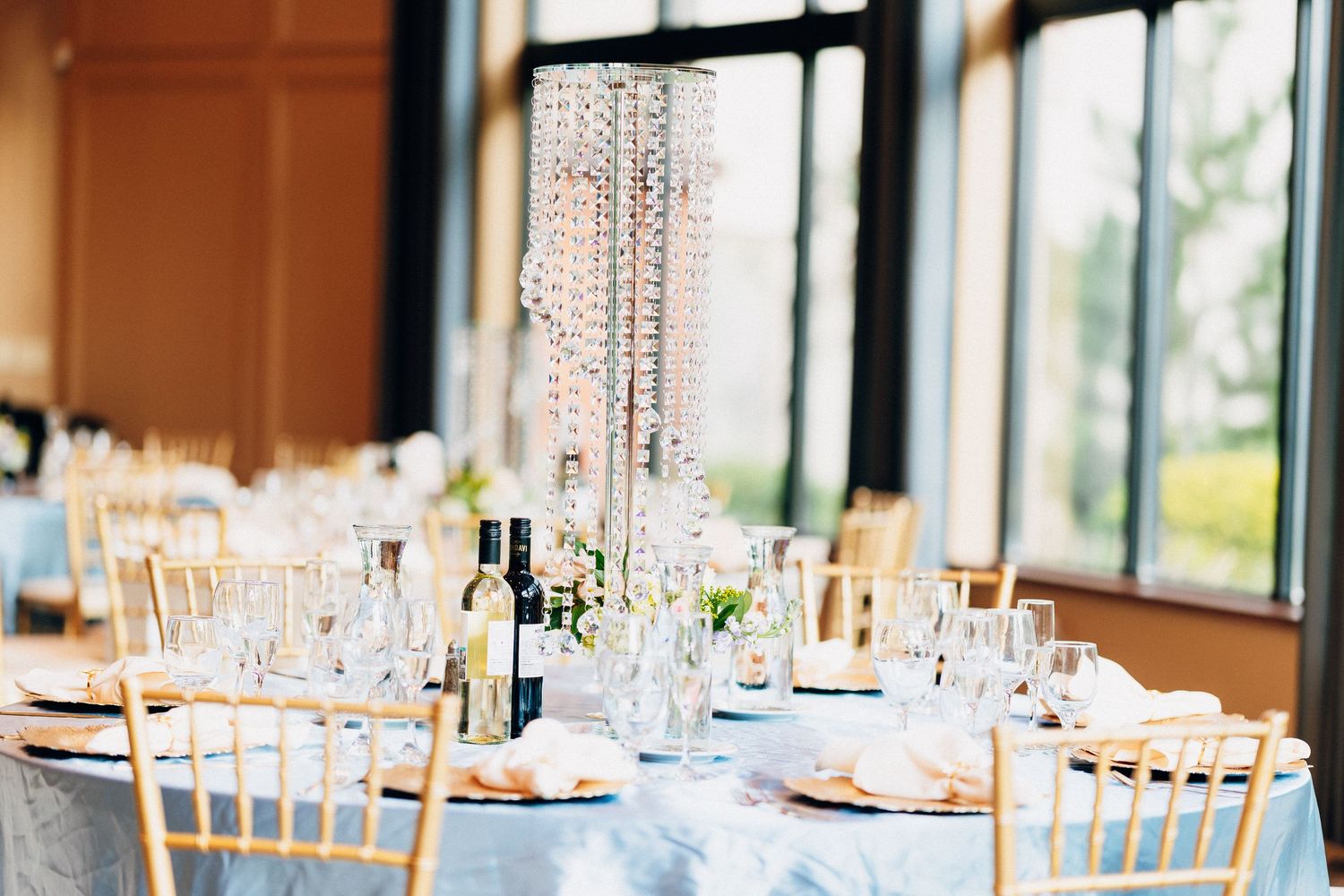 Elegant wedding reception table with crystal chandelier centerpiece, gold chiavari chairs, and blue linen tablecloth.
