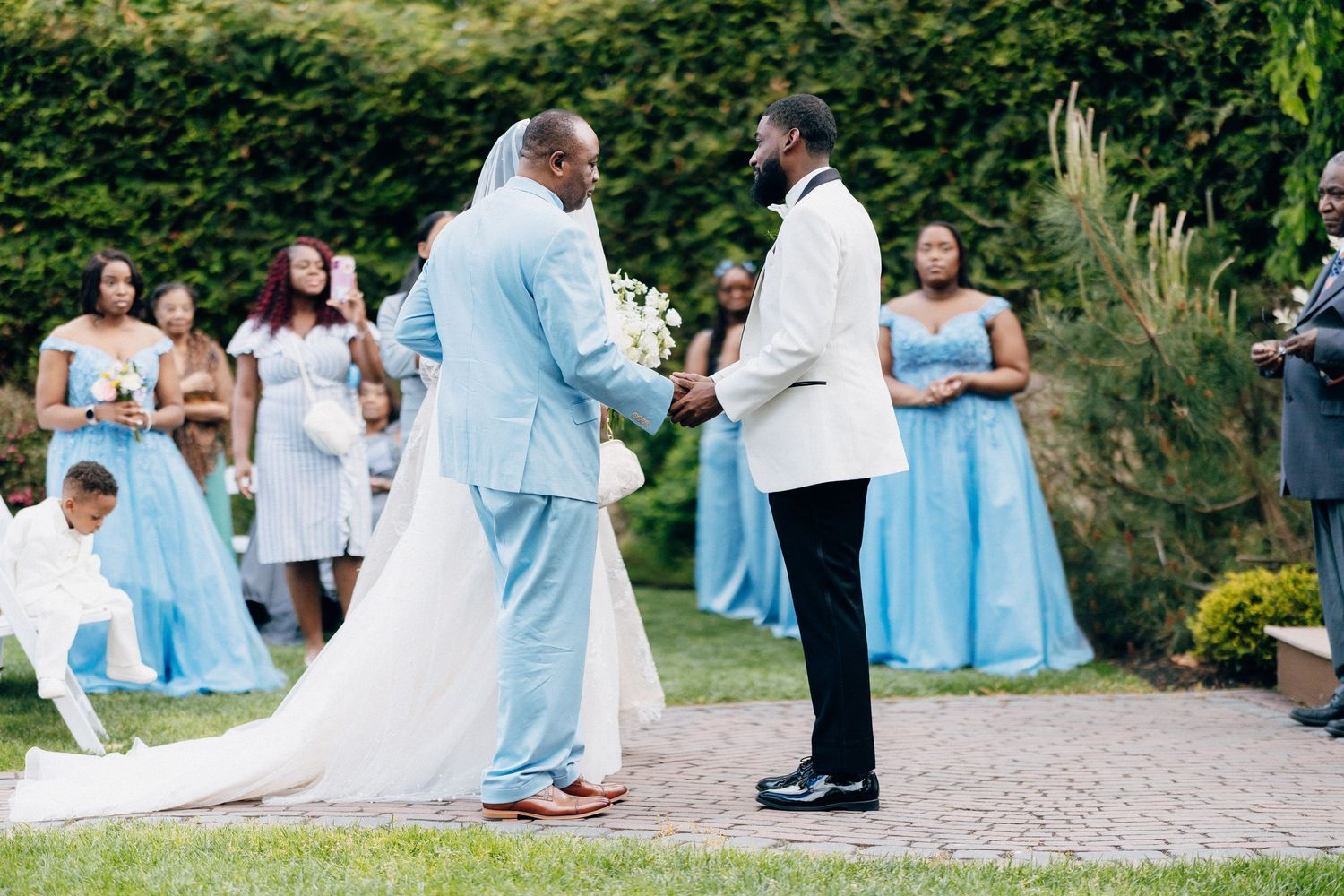 Outdoor wedding ceremony with groom in white jacket, bride in white gown, and bridesmaids in light blue dresses.