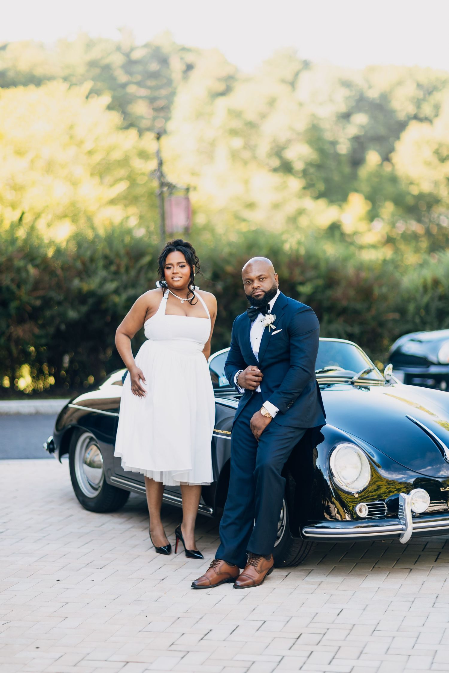 Elegant couple in wedding attire posing beside a classic black vintage car on a brick driveway.