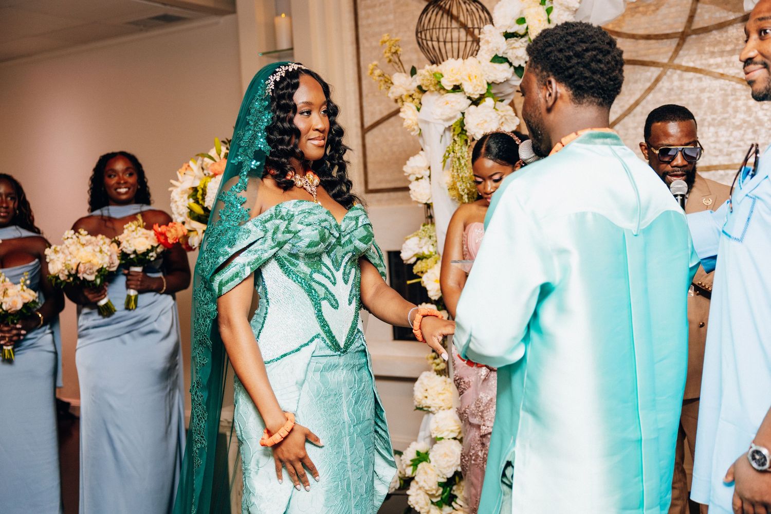 Bride in green lace gown and veil holds groom's hands during wedding ceremony with bridesmaids in blue.