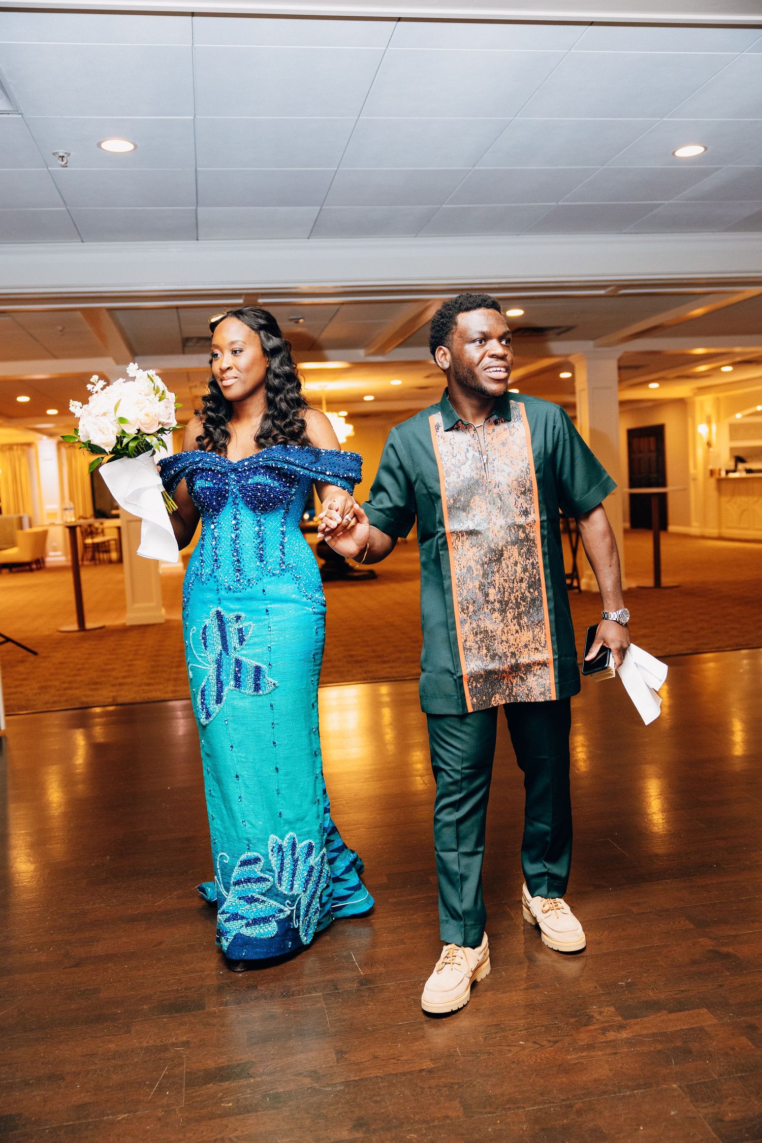 A couple in vibrant African traditional attire walks through an elegant event hall, woman holding white bouquet.