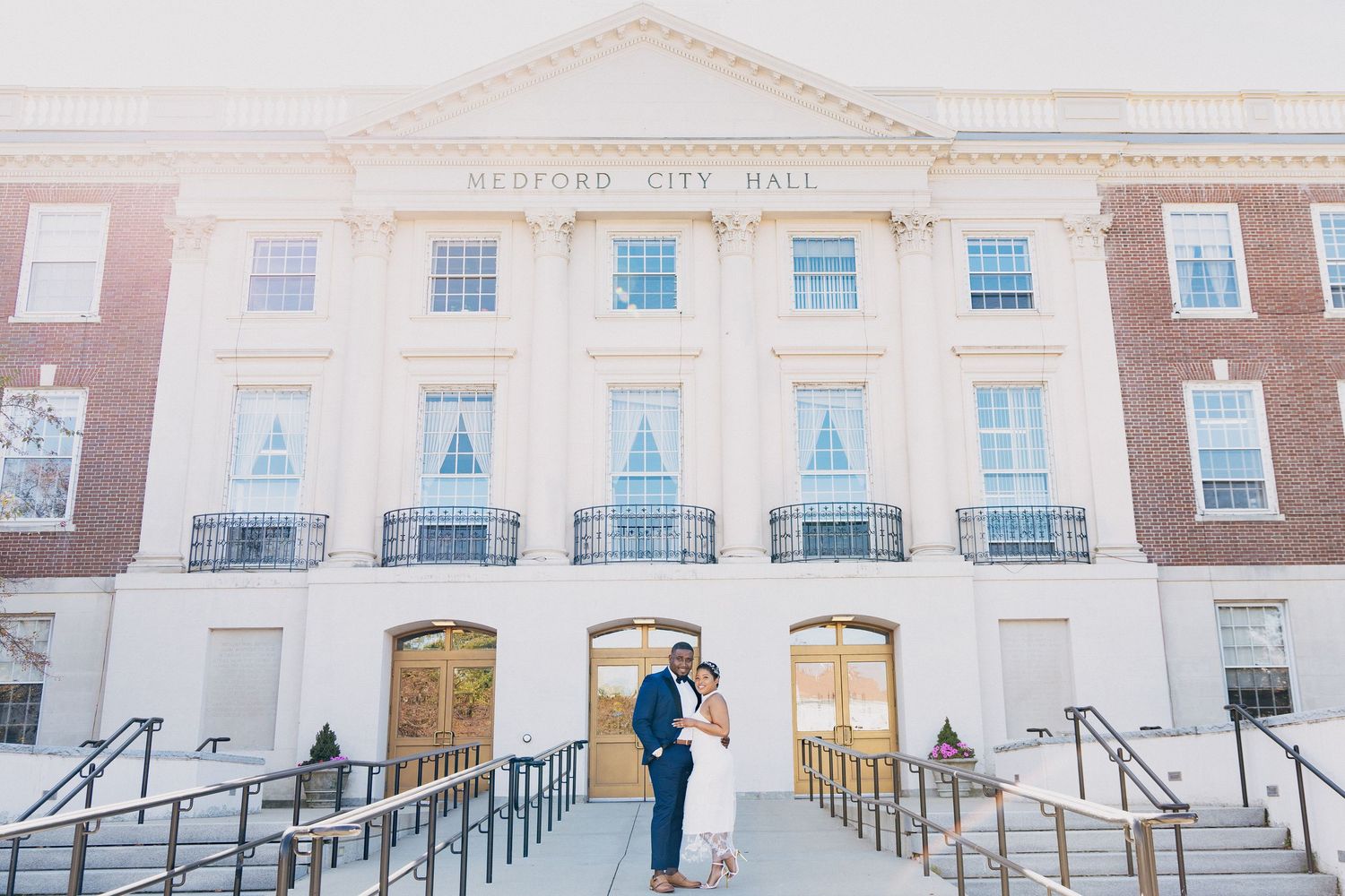 Couple poses on steps of Medford City Hall in wedding attire, brick building with white columns backdrop.