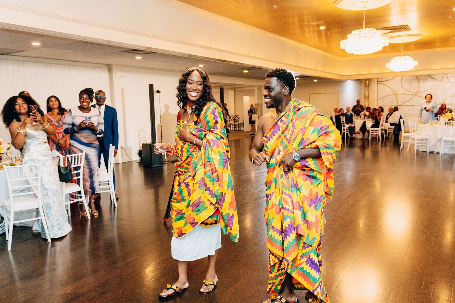 Couple in colorful Kente cloth attire dancing joyfully at a wedding reception in an elegant ballroom.