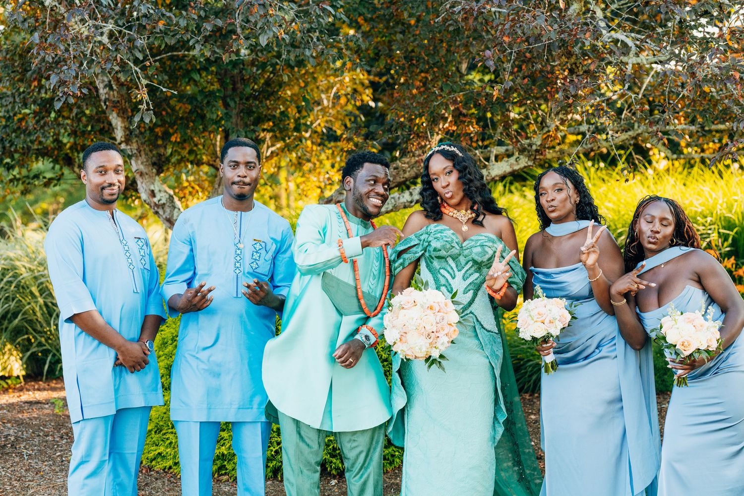 Wedding party in teal and blue outfits posing outdoors with bouquets among autumn trees.