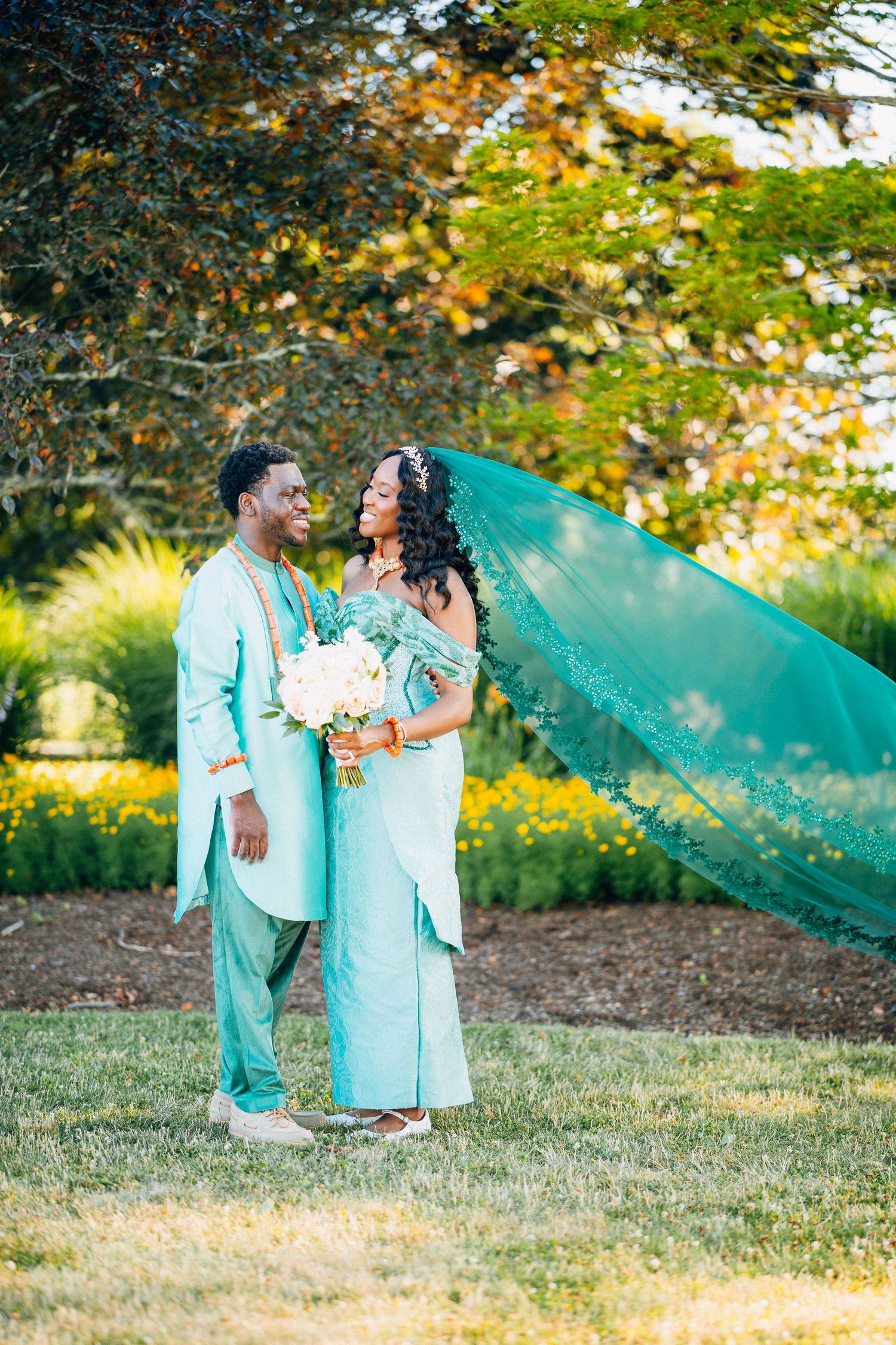 Couple in matching teal outfits pose outdoors, bride holds bouquet with veil flowing in wind.