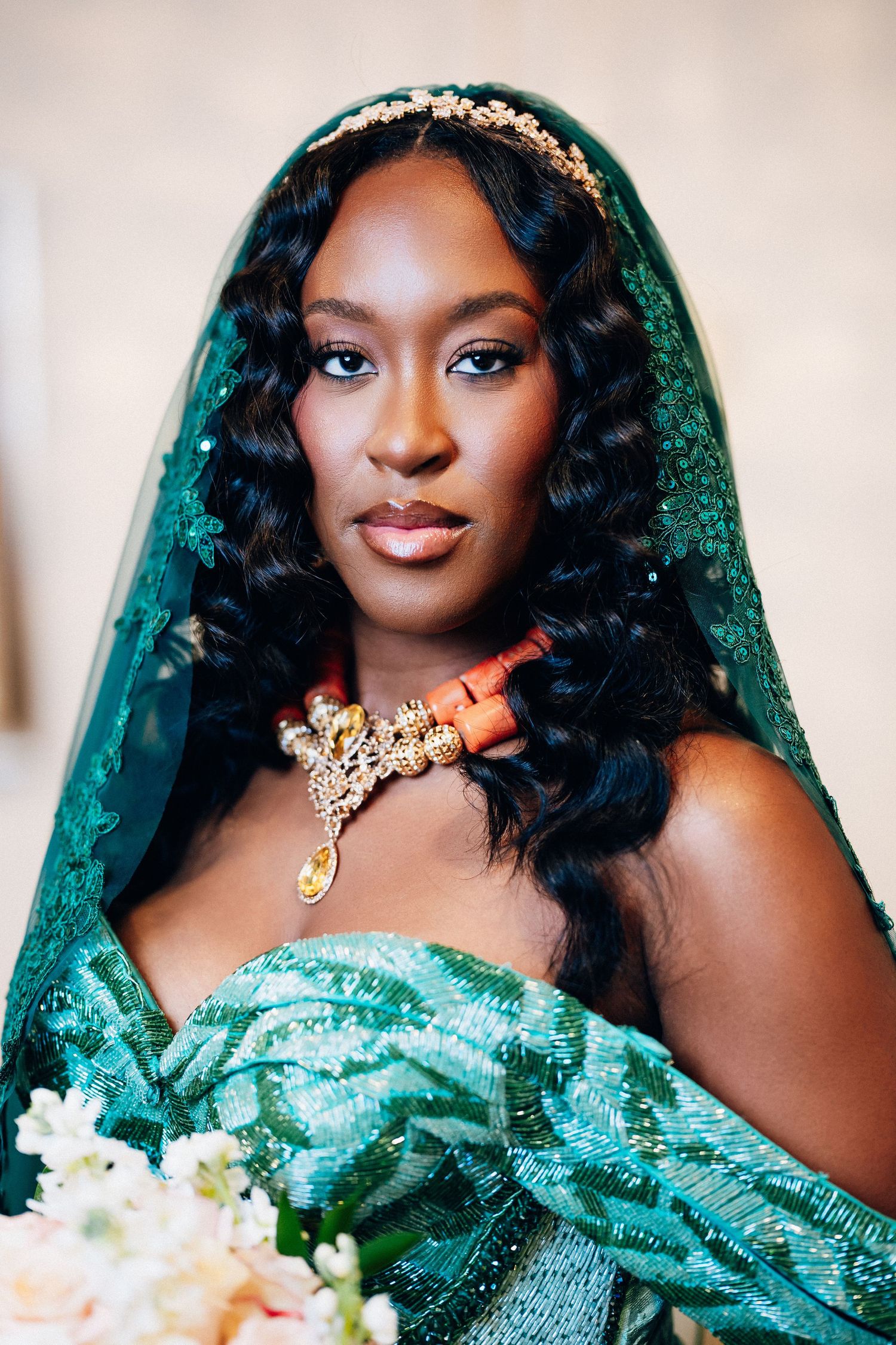 A bride in a green embroidered gown and veil with coral necklace and gold headpiece holds a white bouquet.
