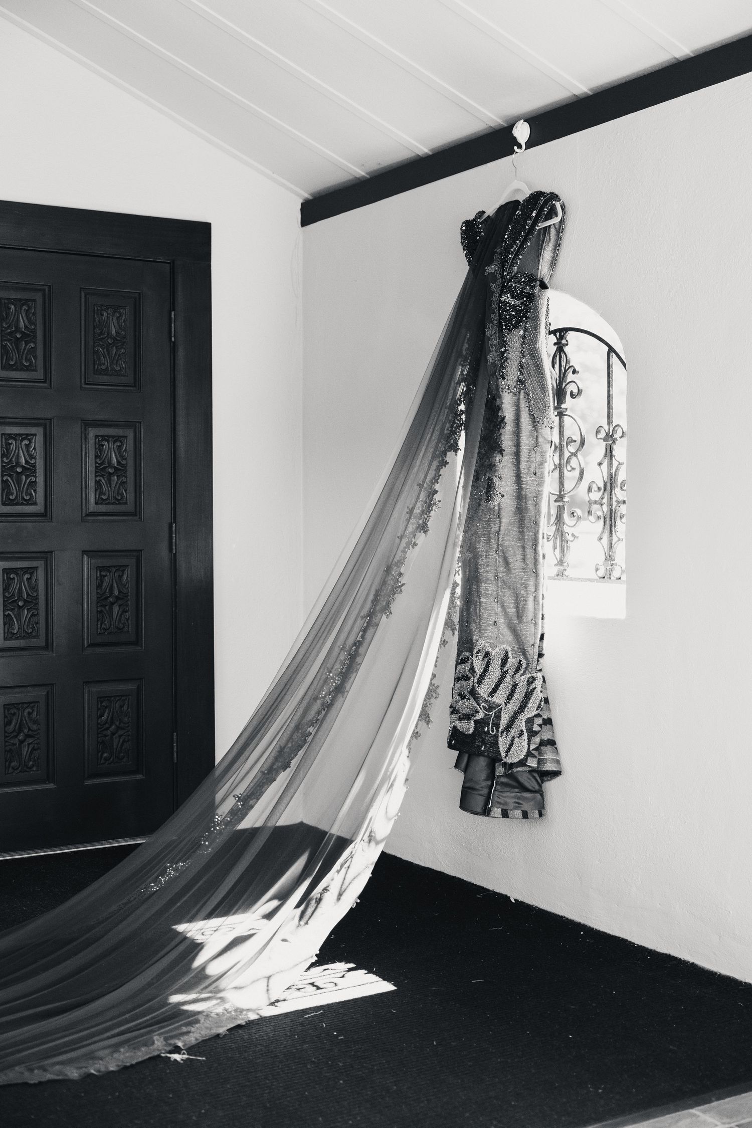 Black and white photo of elegant gown with long train hanging near ornate dark wooden door.