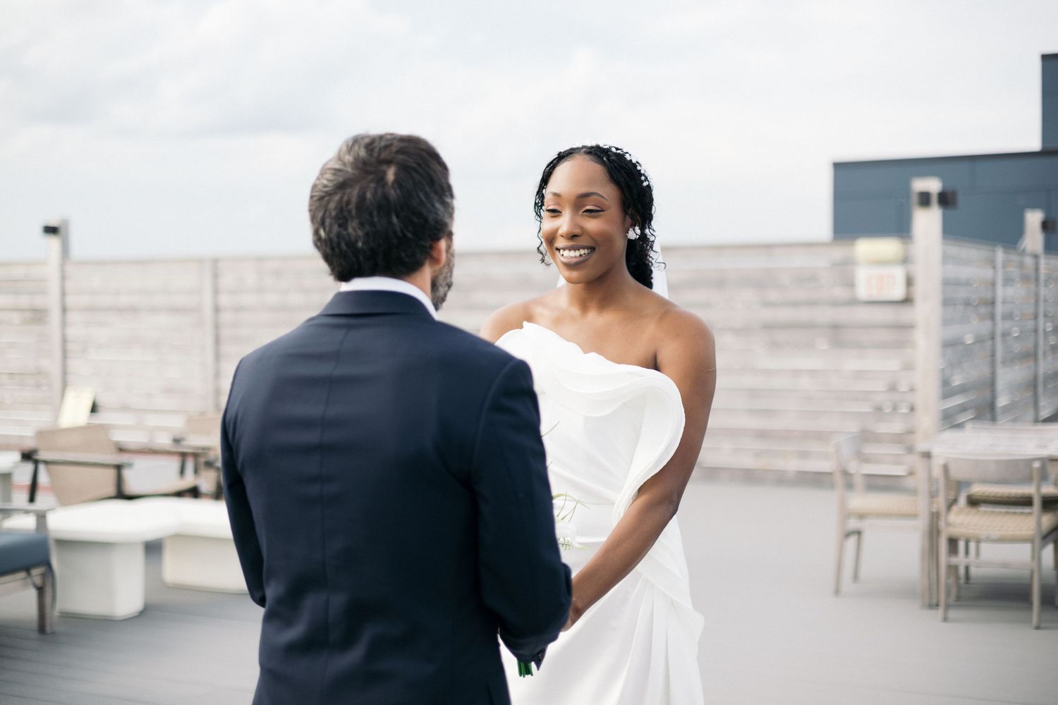 A smiling bride in a white strapless gown holds hands with a groom in a navy suit on a rooftop venue.