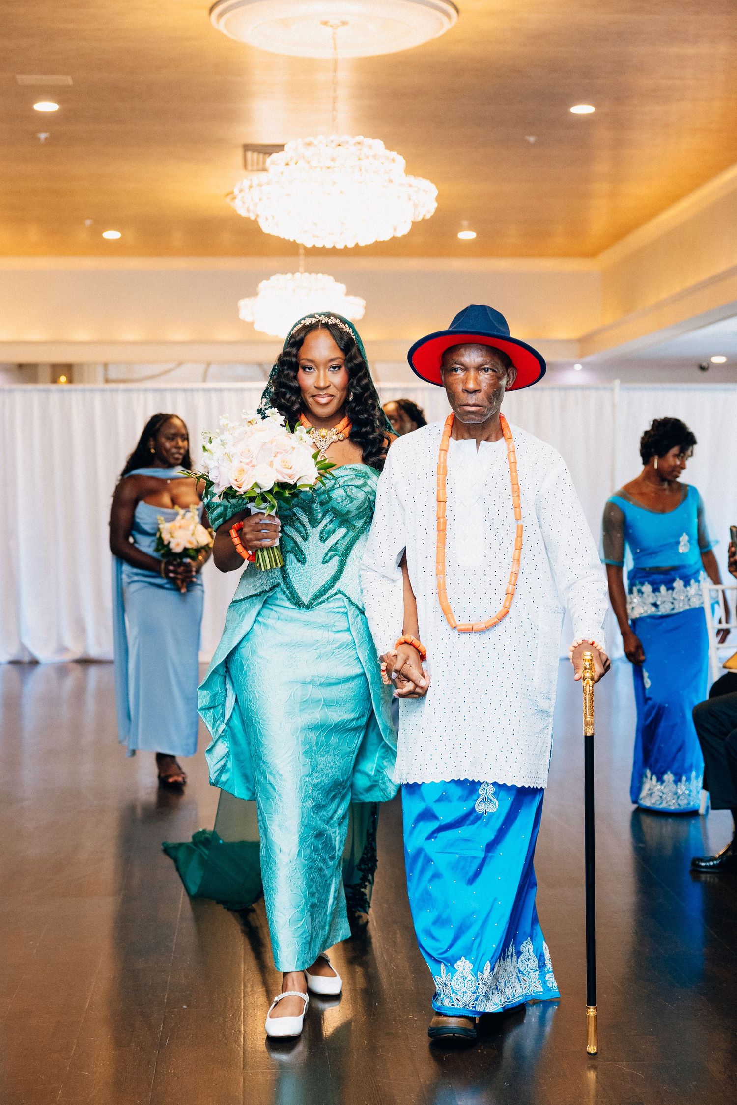 Smiling bride in teal African gown walks down aisle escorted by elder in white and blue traditional Nigerian attire.