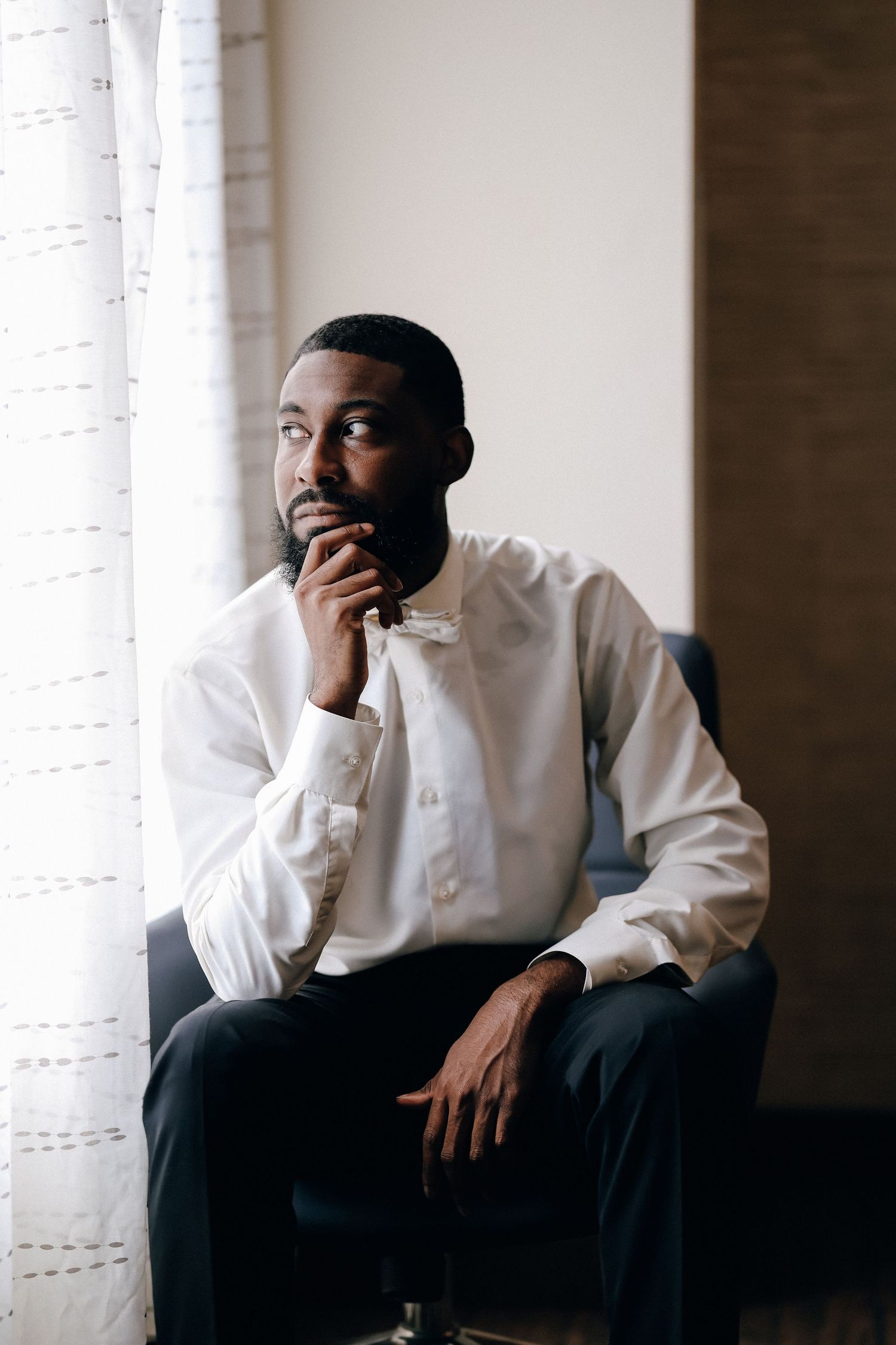 A thoughtful Black man in a white dress shirt sits in a chair by a window, resting his chin on his hand.